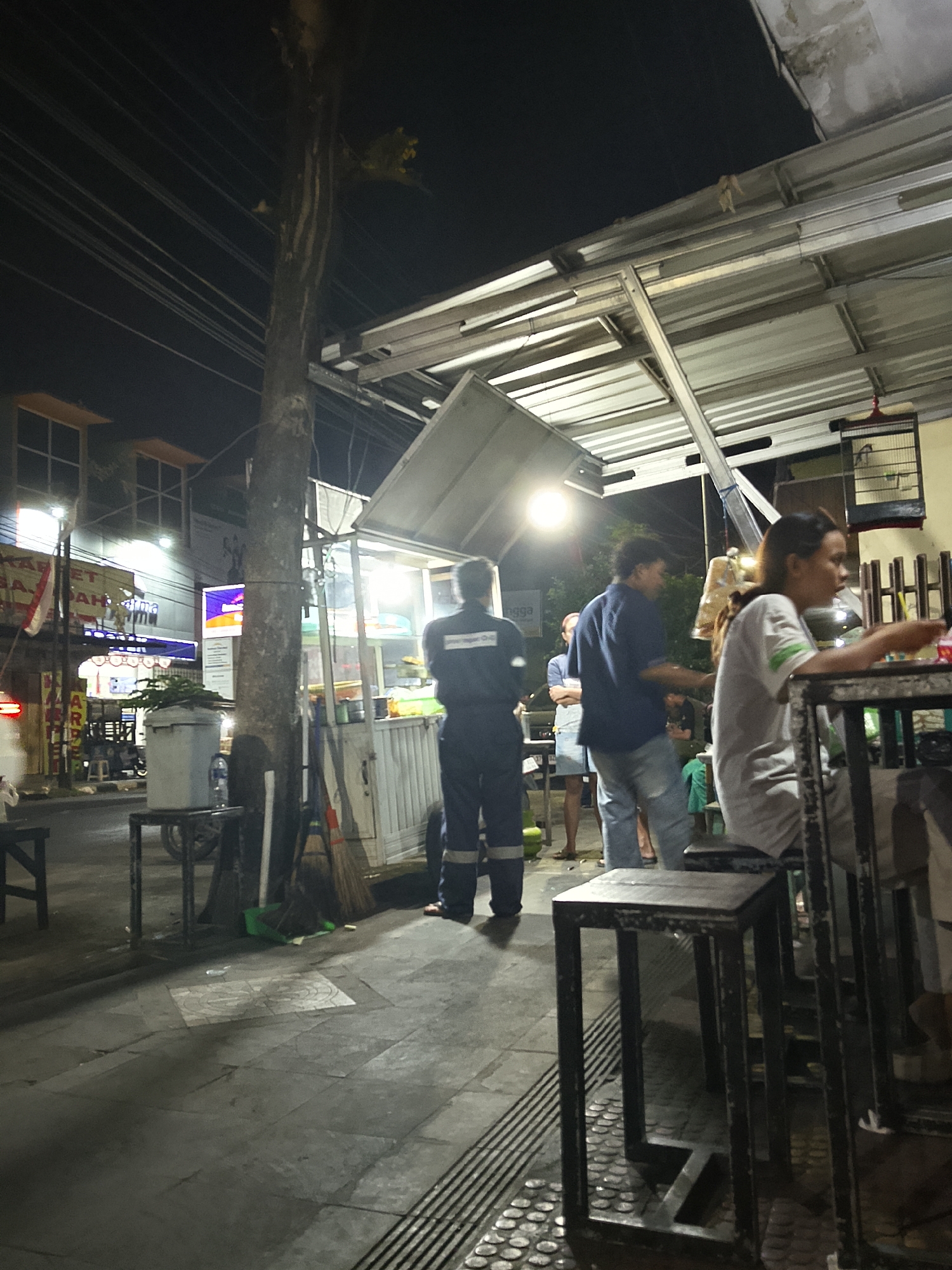 The image shows a street scene at night in Balun, Indonesia, likely a food stall or small restaurant area. The foreground features a dark gray tiled sidewalk with a metal grate, and a black metal stool. To the left, a dark wooden table leg is visible. In the midground, a large tree trunk stands to the left of a food stall with a white awning and metal frame. Inside the stall, food items are visible, illuminated by bright lights. A person in a dark uniform is standing near the stall, facing away from the camera. To their right, another person in a blue shirt and light-colored pants is interacting with someone else. A woman in a white shirt is seated at a table to the right, looking to the side. In the background, there are buildings with lit windows, streetlights, and electrical wires overhead. A red and white flag hangs from a pole on a building to the left. A birdcage is visible hanging from the awning on the right side of the frame. The overall atmosphere is dim and active with people engaged in various activities.