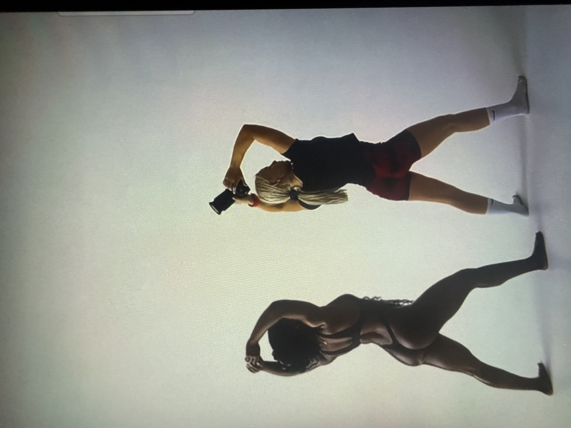 The image depicts two female individuals in what appears to be a photography studio, characterized by a seamless, light-gray backdrop and floor. The scene provides no visual identifiers connecting it to the city of Varna, Bulgaria.
On the left, an individual with dark skin and dark hair, wearing a dark thong-style swimsuit or leotard, stands facing away from the viewer. Her arms are raised and bent, with hands clasped together above her head. Her posture suggests a deliberate pose.
On the right, an individual with lighter skin and blonde hair tied in a ponytail wears a black short-sleeved top, dark red shorts, and white ankle socks. She holds a black camera with a lens extended, positioned above her head. Her head is turned slightly to the right, and her arms are raised and bent as if preparing to photograph or simulating the action of taking a picture. The interaction implies a photoshoot, with the individual on the left posing and the individual on the right acting as a photographer.
The lighting is even and diffused, consistent with an indoor studio setting.
