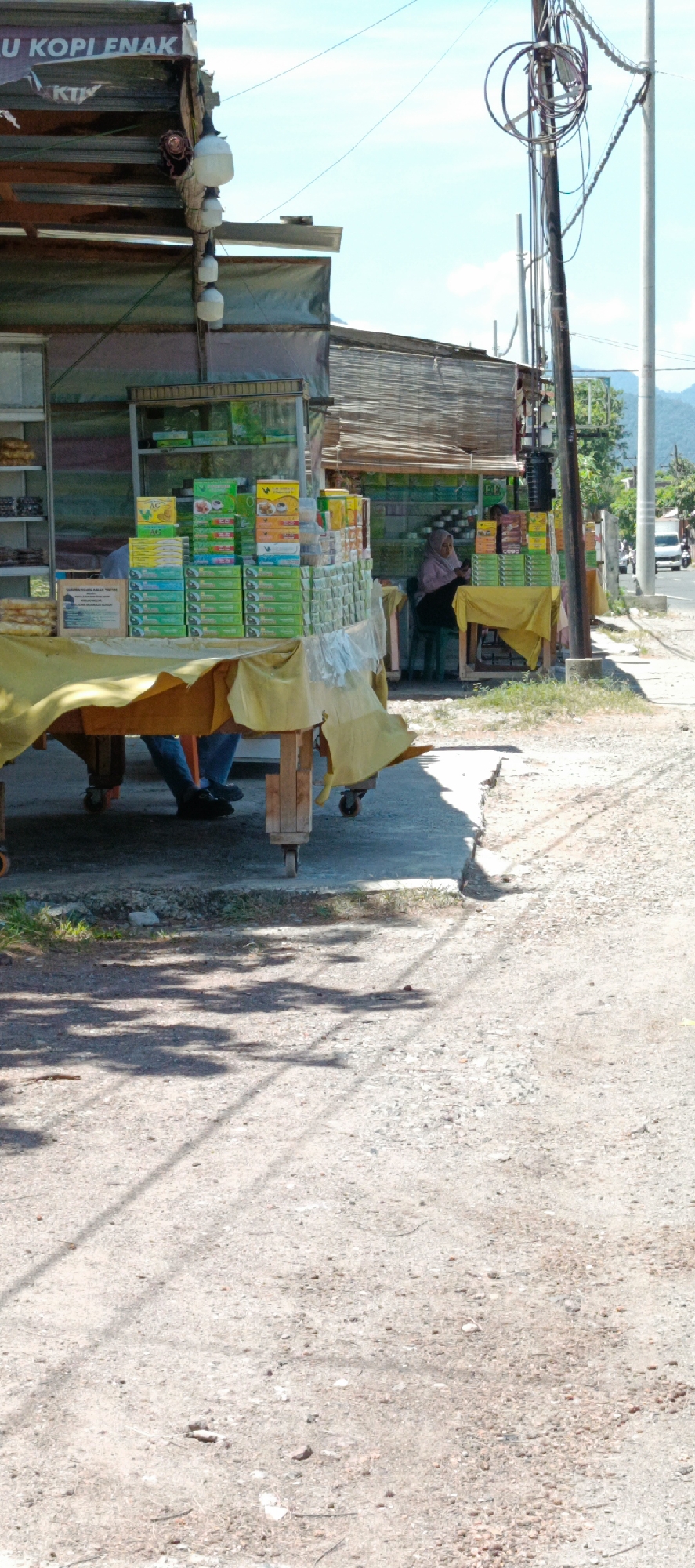 The image depicts an outdoor marketplace stall in Lampisang, Indonesia, featuring a variety of goods, primarily packaged food items. The stall is set up along a roadside with a clear blue sky and distant mountains.
In the foreground, a wooden table covered with a yellow cloth displays numerous stacked boxes of food products, predominantly in green and yellow packaging. Some of these boxes appear to be snacks or confectionery. Two individuals are partially visible under the table; one appears to be wearing blue jeans and dark shoes, while the other's lower body is also visible.
Behind the table, the main stall structure is visible, made of corrugated metal roofing and wooden supports. Hanging from the roof are several industrial-style light fixtures. Further back, a glass display case is filled with more packaged goods. Another person, a woman wearing a headscarf, is seated behind a smaller stall or table, interacting with the products. She is dressed in a pink hijab and dark clothing.
The environment suggests a warm, sunny day. The ground in front of the stall is a mix of gravel and dirt, with some patches of grass. Shadows from overhead trees are cast across the ground. In the background, a utility pole with numerous wires stands tall. The road adjacent to the stall shows some vehicular traffic, indicating it is an active thoroughfare. The overall scene is that of a typical roadside market in a rural or semi-urban Indonesian setting.