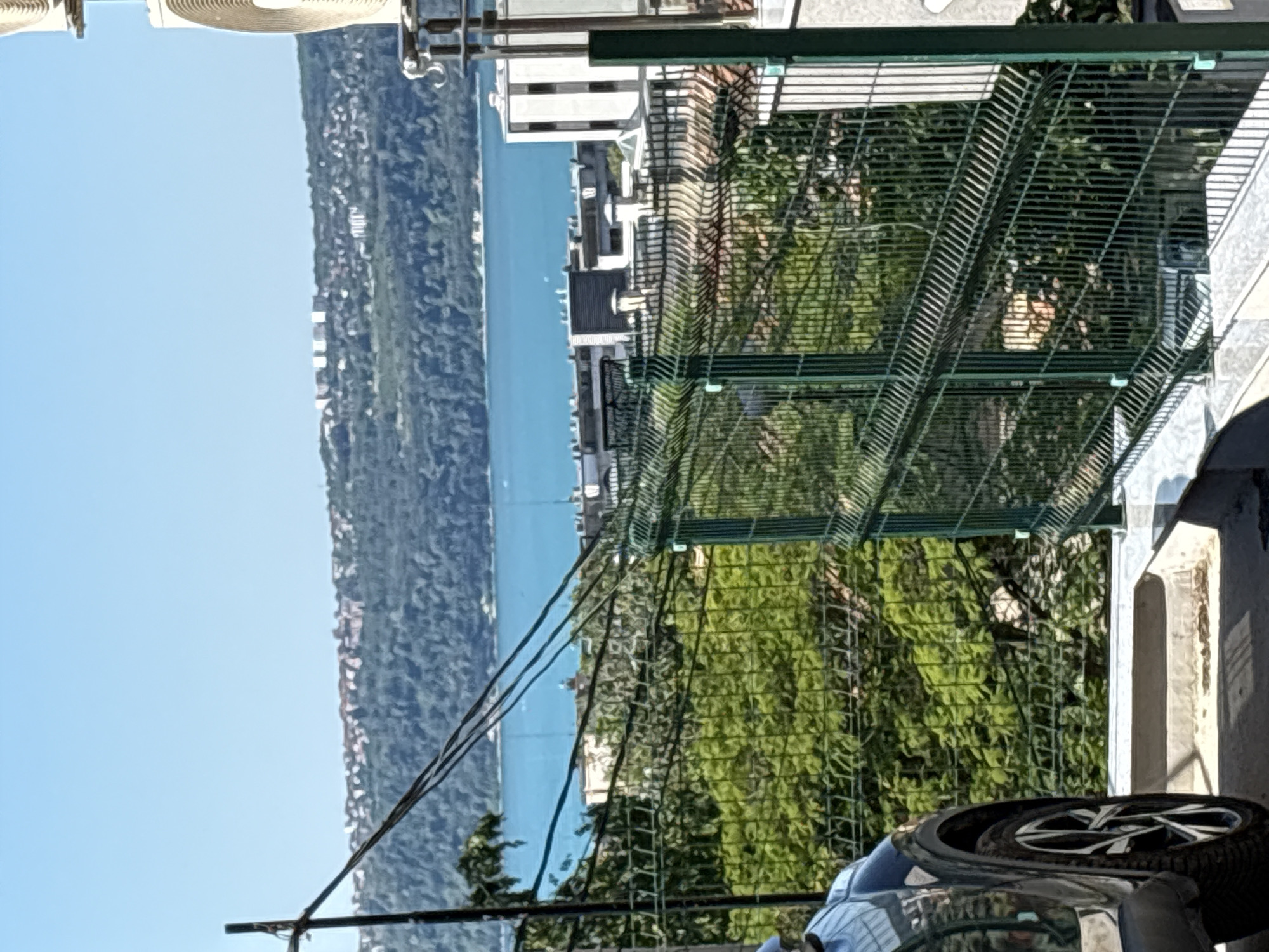 The image captures a scenic view of the Black Sea from a hillside location in Varna, Bulgaria. In the foreground, a dark-colored car is partially visible on the left, with its wheel and fender in focus. A green metal fence stretches across the mid-ground, creating a barrier between the viewer and the landscape beyond. Behind the fence, lush green trees form a dense foliage.
Further in the distance, the vibrant turquoise waters of the Black Sea are clearly visible, meeting a distant shoreline characterized by a hilly terrain covered in trees and dotted with buildings. Several modern structures, including apartment buildings with white facades and dark windows, are scattered across the hills. The sky above is a clear, bright blue, indicating a sunny day. Electrical wires are strung across the upper portion of the image, supported by a dark pole on the left. The overall impression is one of a pleasant, urban environment with a prominent natural landscape.
