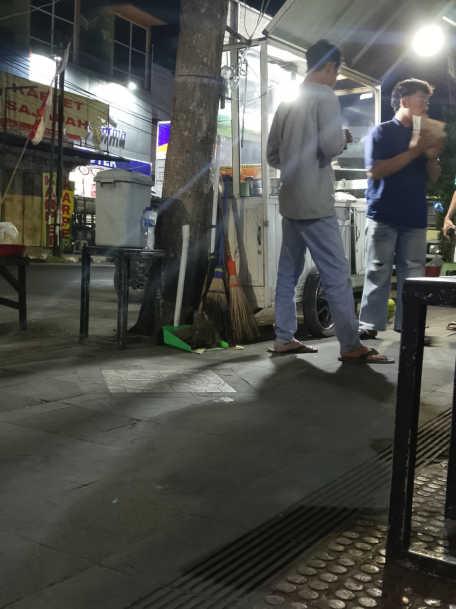 In Balun, Indonesia, at night, two individuals stand near a street food cart. The person on the left, wearing a gray long-sleeved shirt and light blue pants, is holding a small item. The person on the right, in a blue t-shirt and ripped light blue jeans, holds a paper container. A large tree trunk is prominent in the foreground. Various objects are visible: brooms leaning against the tree, a green dustpan with debris, a trash bin, and a cooler on a metal stand. The ground is paved with tiles and shows reflections from the overhead lighting of the food cart. Signs with text, including "KARPET SAJADAH," are present in the background.