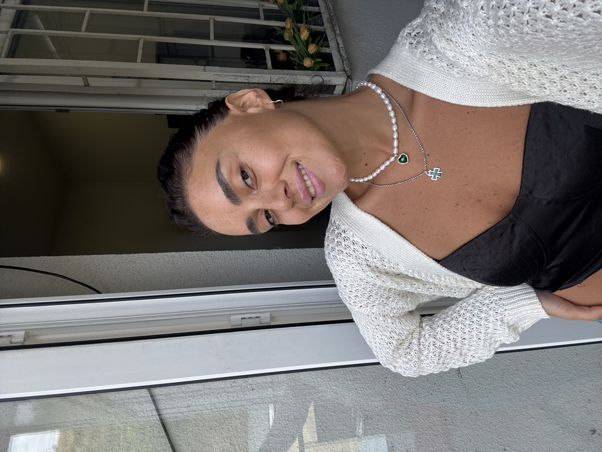 A woman with dark hair and light skin is taking a selfie in front of a window. She is wearing a white knitted cardigan over a black top. She has on a pearl necklace with a green heart-shaped pendant and another silver chain with a cross pendant. She is smiling at the camera. The window has a white frame and a security grate. Outside the window, there are yellow flowers. The location is likely a residential building in Tbilisi, Georgia, given the architectural style of the window and the stucco texture of the wall.