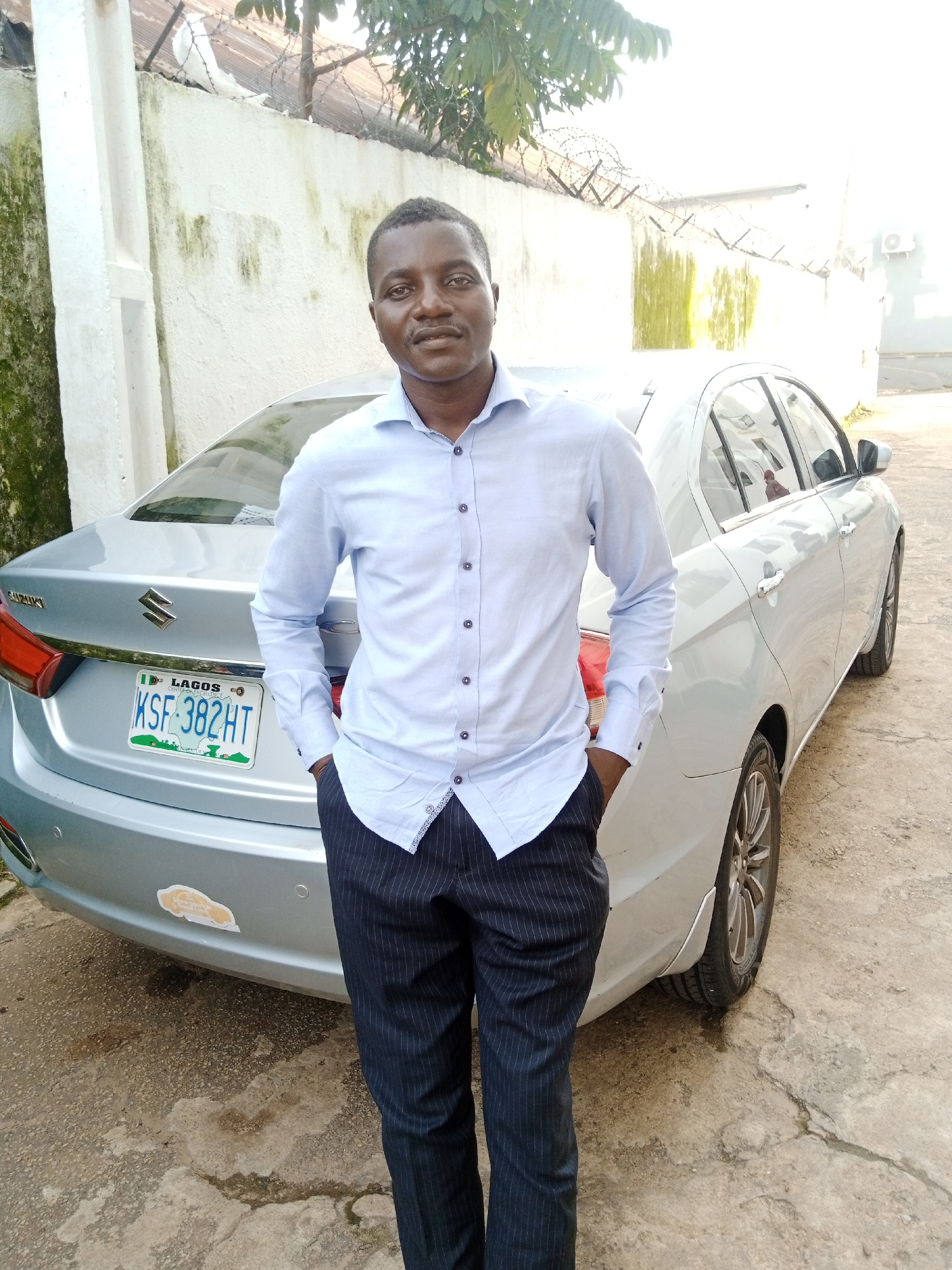 A medium-shot portrait shows a Black man standing in front of a silver sedan. He is wearing a light blue collared shirt with dark pinstripe trousers and his hands are in his pockets. The car is parked on a concrete surface, and a white wall with green moss stains and barbed wire along the top is visible behind the man and the car. The license plate of the car reads "KSF 382HT" and displays "LAGOS". The scene appears to be an outdoor location in a city, potentially Jos, Nigeria, given the context provided. The lighting suggests it is daytime, with bright sunlight casting shadows.