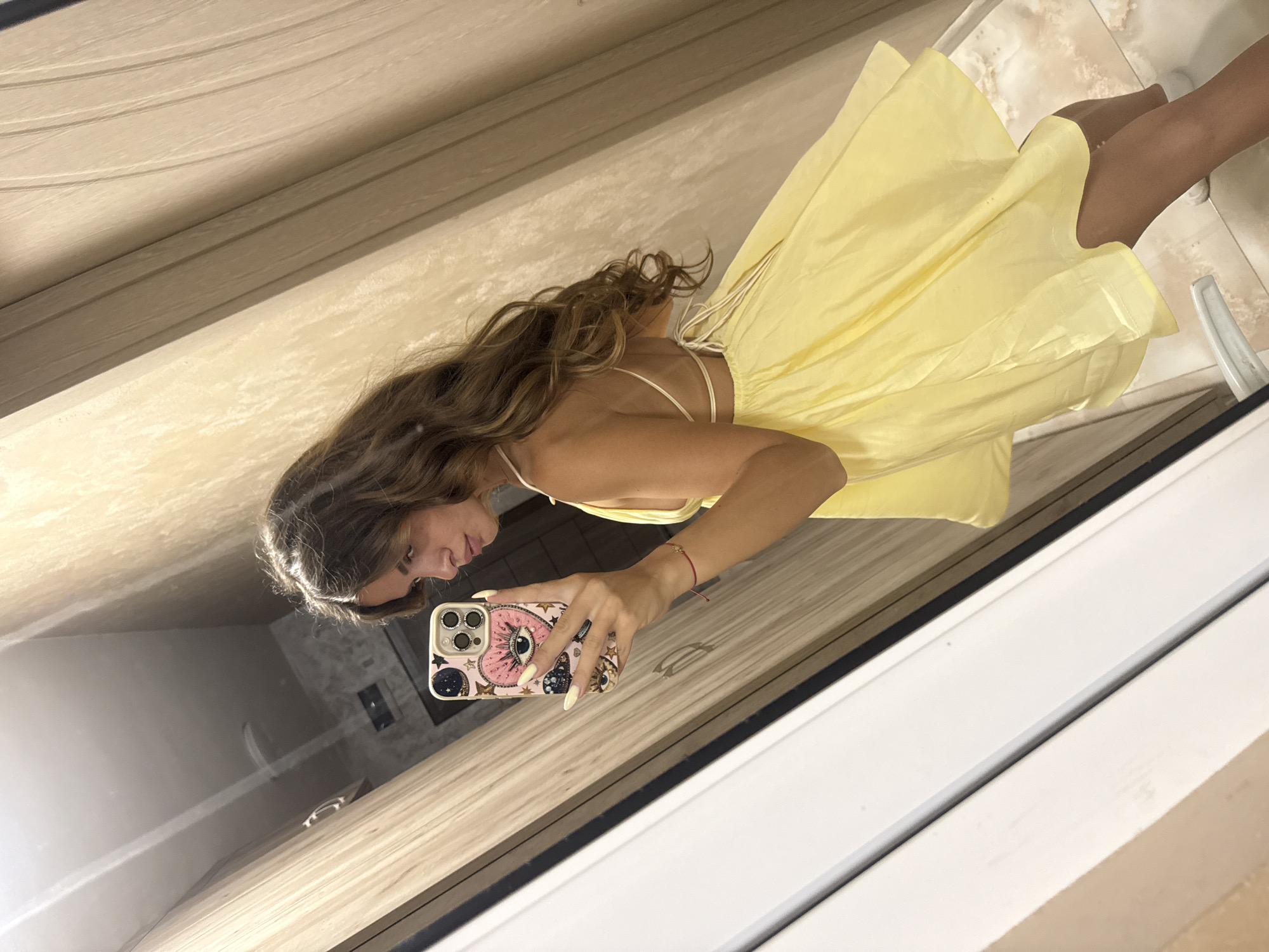 A young woman with long, wavy brown hair takes a mirror selfie in a hallway with wood-paneled walls and white trim. She is wearing a bright yellow, spaghetti-strap dress. The dress has a low back with a tie closure. She is holding a smartphone with a patterned case that features an eye motif. She has long, light-colored fingernails and a red string bracelet on her left wrist. The floor appears to be tiled. The scene is not identifiable as being in Plovdiv, Bulgaria.