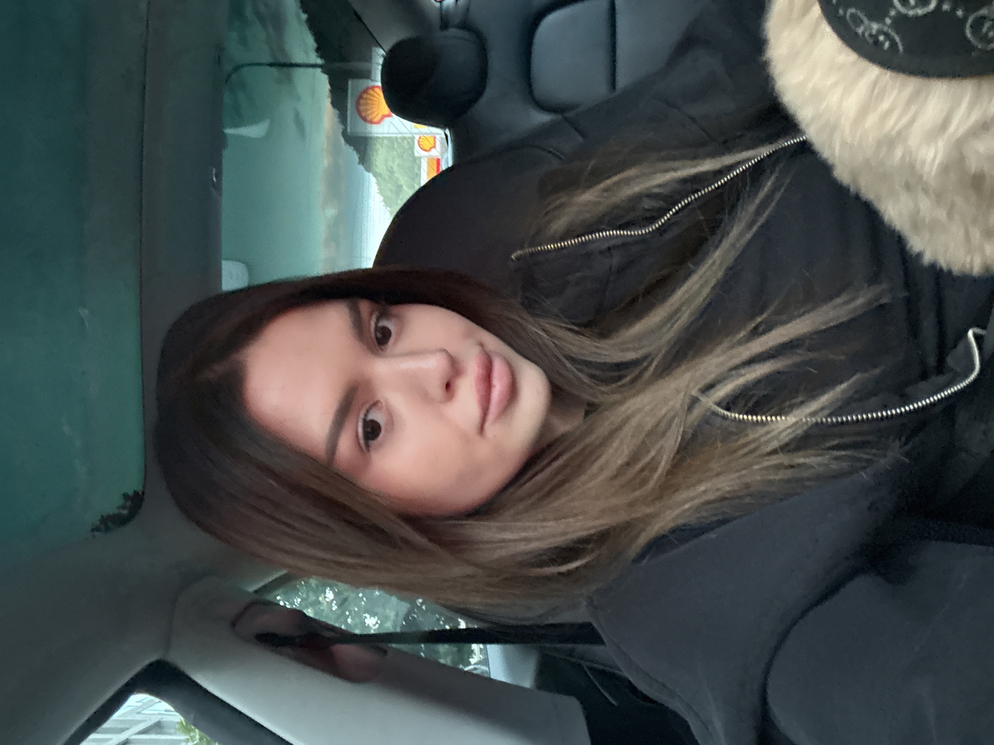 A woman with brown hair is seated in the driver's seat of a car, with her face angled towards the camera. She is wearing a black zip-up hoodie and appears to be holding a fluffy, beige object in her lap. The interior of the car is visible, including black leather seats and a panoramic glass roof. Outside the car, through the windshield, two Shell gas station signs are visible against a backdrop of trees and a body of water, with a cloudy sky overhead. The scene appears to be set in a city environment, with buildings visible to the left of the frame.