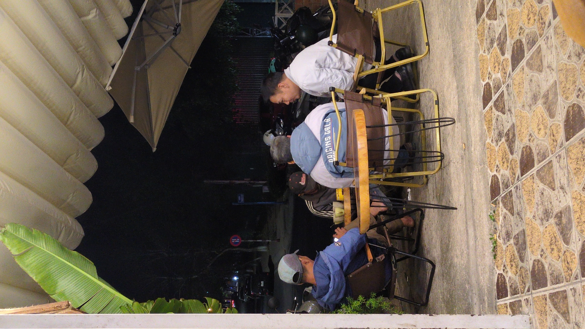 Four men are sitting at a low table in an outdoor cafe in Da Lat, Vietnam, at night. The cafe has tile flooring and is located on a sidewalk. The men are sitting in metal-framed chairs with brown leather or vinyl seats and backs. The man closest to the camera is wearing a blue jacket and a baseball cap. The man next to him is wearing a black Adidas jacket. The next man is wearing a denim jacket with "ORIGINS GALA" printed on the back. The fourth man is wearing a white jacket. They appear to be talking or looking at something on the table. The background is dark, but there are streetlights and some vehicles visible. There is a large banana leaf and a partially deflated white inflatable structure in the foreground. A large beige umbrella is also present. There are motorbikes parked nearby.