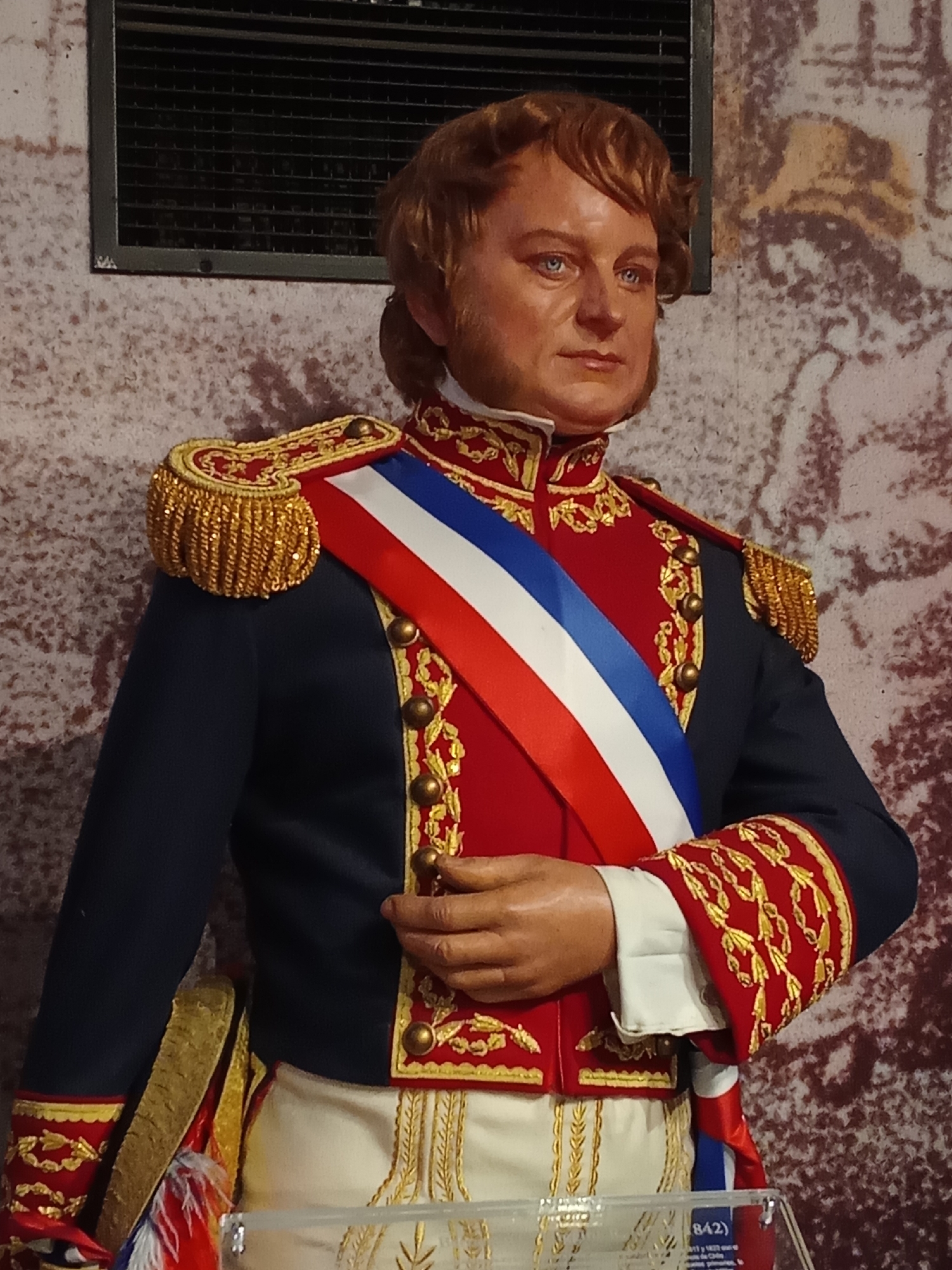 The image shows a life-sized wax figure of a man in a historical military uniform. The figure is positioned in front of a textured backdrop that appears to be a wall with a faded mural or tapestry. The man has light brown hair styled in a somewhat disheveled manner, and his gaze is directed slightly to the right. He is wearing a dark blue military jacket adorned with red lapels and elaborate gold embroidery. A sash in the colors of red, white, and blue is draped diagonally across his chest. His lower garment appears to be white breeches with gold trim. He has a white cravat at his neck and ornate gold epaulets on his shoulders. The figure's right hand is slightly raised, with his fingers curled. In the lower right corner of the image, a partial view of a glass display case with text and a number "842)" is visible. This figure is part of an exhibition at the Museo Histórico Nacional in Santiago, Chile, depicting historical figures relevant to Chilean history.