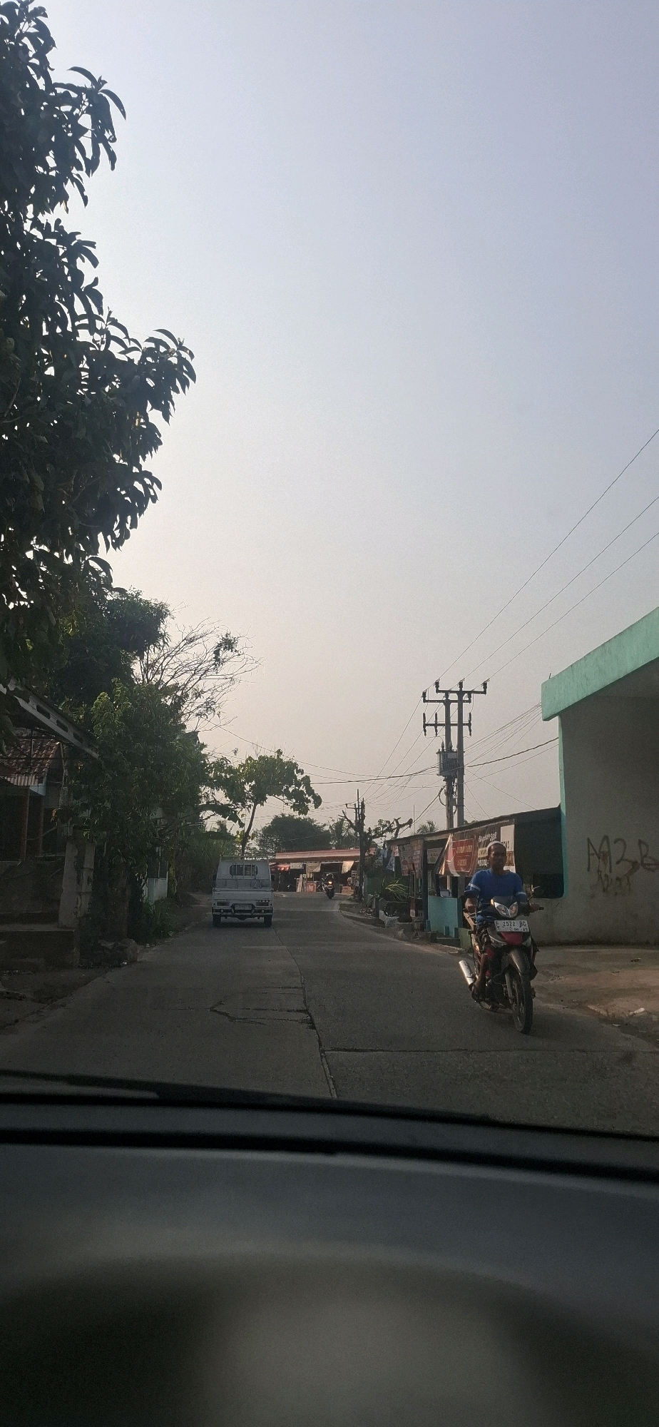 The image depicts a view from inside a vehicle, looking out onto a street in Cibinong Hilir, Indonesia. The sky is overcast and pale. On the left side of the street, there are houses and trees. A white pickup truck is parked on the road. Further down the street, a man is riding a motorcycle towards the camera. He is wearing a blue shirt and dark pants. The road is asphalt and appears to be in moderate condition with visible cracks. Utility poles with wires are visible along the street. On the right side of the street, there is a building with a green roof and graffiti on its wall. The overall scene suggests a typical day on a local street in Indonesia.
