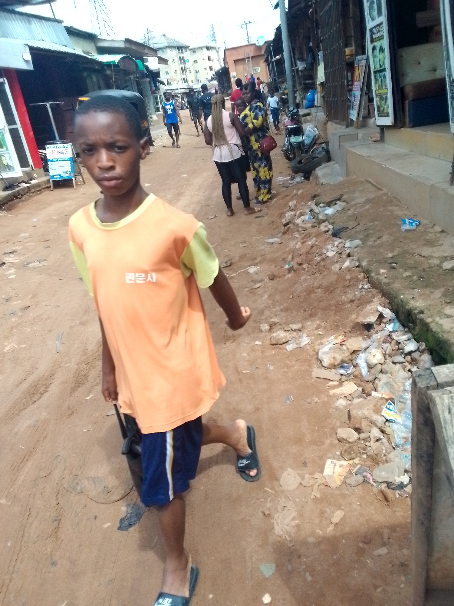 A young boy walks down a dirt road in Nkpor, Nigeria. He is wearing an orange t-shirt with yellow sleeves and dark blue shorts. His t-shirt has Korean writing on it. He is wearing black flip-flops. The road is lined with small shops and buildings. There is trash on the side of the road. In the background, other people are walking and standing around. One woman is wearing a colorful patterned dress. A motorcycle is parked on the side of the road.