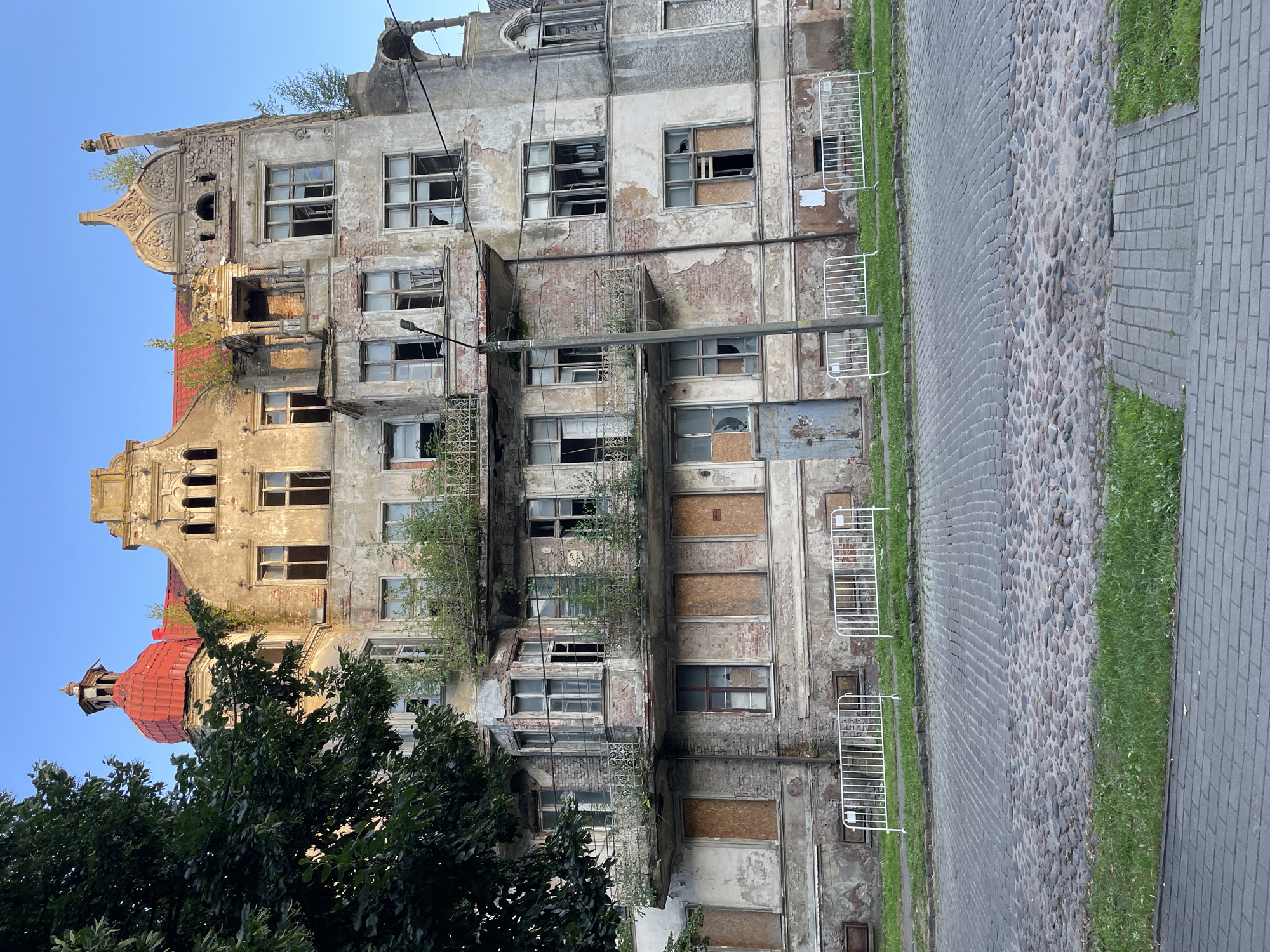 The image shows a large, dilapidated building on a cobblestone street in Sovetsk, Russia. The building appears to be abandoned, with boarded-up windows and peeling plaster. There are several balconies with decorative iron railings, some of which are overgrown with vegetation. The roof is red tile, and there is a red domed turret on the left side of the building. A tall street lamp stands in front of the building. White metal barriers are placed along the front of the building, and grass grows in front of the barriers. The street is paved with cobblestones and a section of grey brick pavement is visible in the foreground. The sky is clear and blue. There are no people visible in the image.