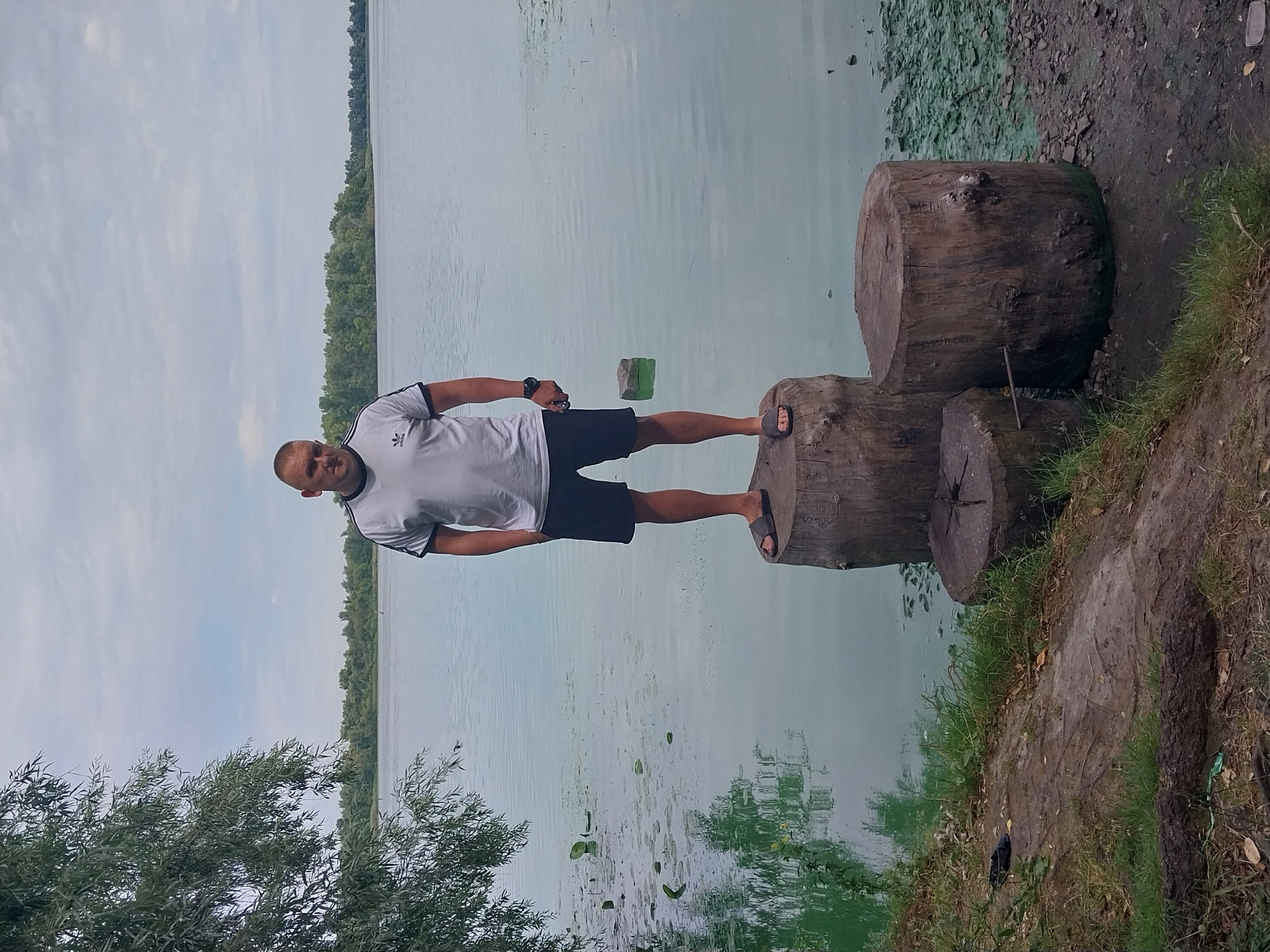 A man stands on a thick wooden stump by a large body of water, possibly a river or lake. He is wearing a white Adidas t-shirt with black stripes on the sleeves and black shorts, along with dark flip-flops. His hands are in his pockets, and he is looking at the camera. The water is calm and appears to have some green algae or plants on the surface, especially near the shore. The background shows a line of trees across the water, and the sky is cloudy. The foreground consists of the shore with grass, dirt, and more wooden stumps. The location is the Dnipro River, with the Prydniprovsk Park visible in the distance on the right bank of the river.