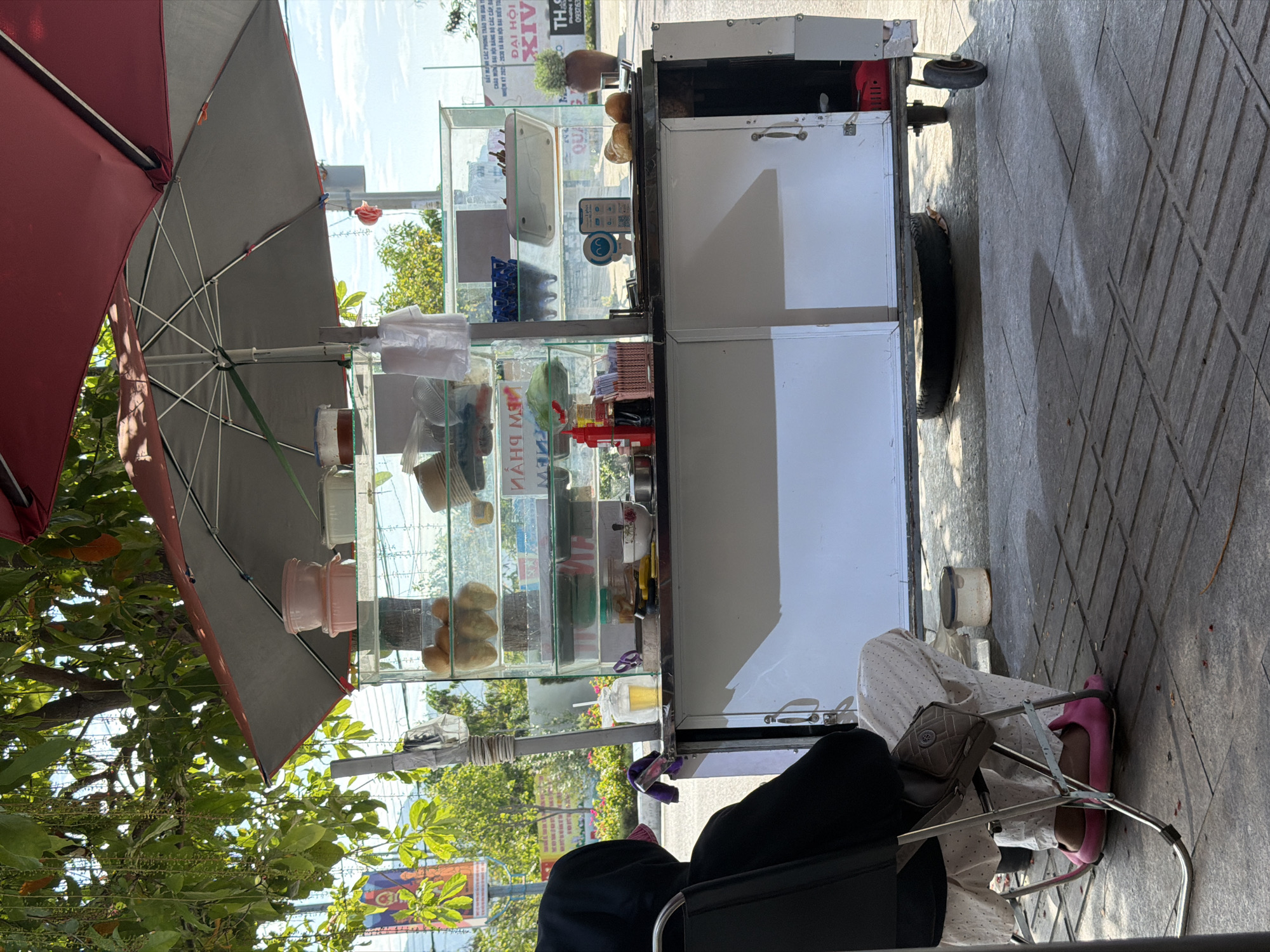 The image captures an outdoor street food scene in Ninh Chu Ward, Vietnam, under bright daylight with distinct shadows indicating mid-day. A mobile food cart, featuring white cabinets and a glass display, is set up on a paved sidewalk. It is shaded by a large umbrella with a grey top and red underside.
The cart's display case contains numerous small bread rolls, likely ingredients for Bánh Mì sandwiches, along with various containers, plastic bags, and utensils. A mobile phone displaying a QR code, presumably for cashless payment, is visible on the cart's counter.
Two individuals are present: a vendor, partially obscured behind the cart and wearing black clothing, and a customer seated on a metal folding chair. The customer is wearing white polka-dotted pants and pink slippers, with a brown crossbody bag resting nearby.
The background shows lush green trees and foliage, with distant buildings and overhead power lines under a clear sky. A large banner or sign in the distance displays Vietnamese text, including "ĐẠI HỘI" (Congress/Conference) and "NHIỆM KỲ 2023 - 2020," suggesting a local public event. Other legible text includes "093763," which appears to be a phone number, and a likely reference to "BÁNH MÌ" (bread/sandwich) on the cart's signage. The scene depicts a common daily transaction at a bustling street food stall.