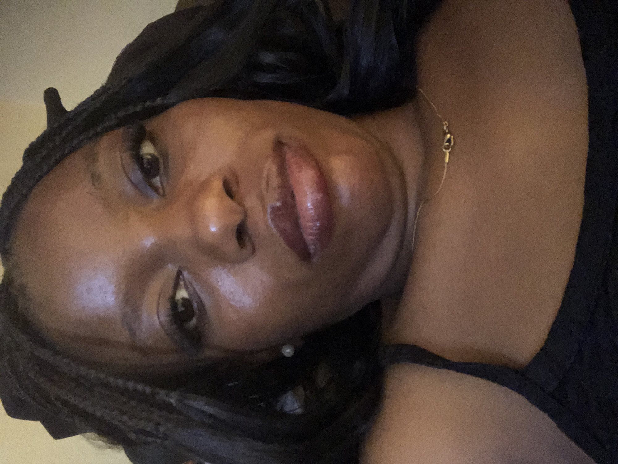 A close-up, medium shot shows the head and upper chest of a Black woman. She is facing forward with her head tilted slightly upwards and to her left. Her dark braided hair is pulled back. She is wearing a black tank top and a thin gold necklace with a clasp. She has on subtle makeup, including eyeliner and lip gloss. Her left earlobe is adorned with a small pearl earring. The background is a blurred beige wall. The scene appears to be indoors.