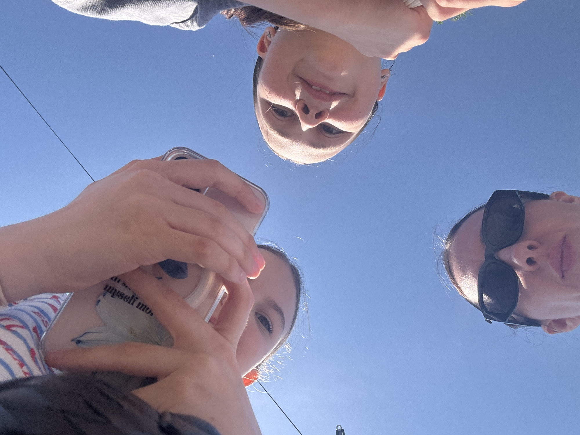 Three young women are positioned in a circle, looking down at the camera. One woman is holding a phone, the case of which has text visible. The background is a clear blue sky. The location is indeterminate; there is no evidence to support the location being Votkinsk, Russia.
