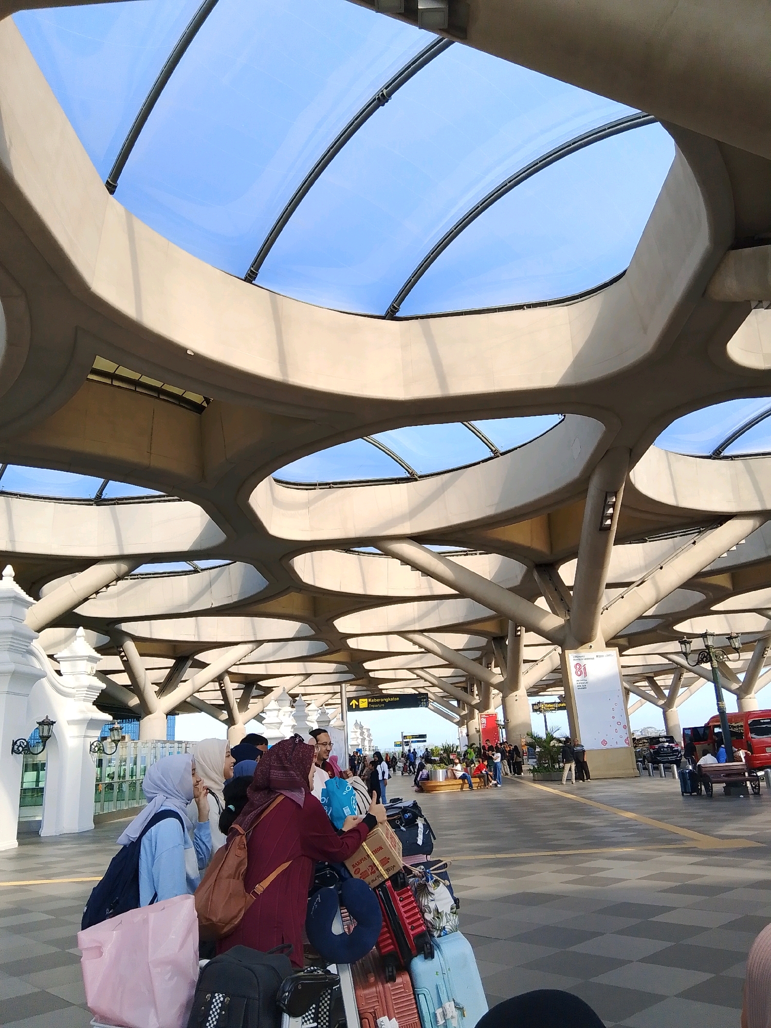 The image depicts the exterior of the Yogyakarta International Airport in Palihan, Indonesia. The distinctive architectural feature of the airport is its honeycomb-like concrete roof structure with translucent panels that allow sunlight to filter through. In the foreground, several people, mostly women wearing hijabs, are gathered with their luggage. They appear to be waiting or in transit, with various suitcases, duffel bags, and shopping bags piled around them. One woman in a maroon hijab is taking a selfie with her phone, while others are looking at their phones or conversing. In the background, there are more travelers, cars, a red bus, and signage indicating "Departure." The overall scene conveys a sense of movement and activity typical of an airport environment.
