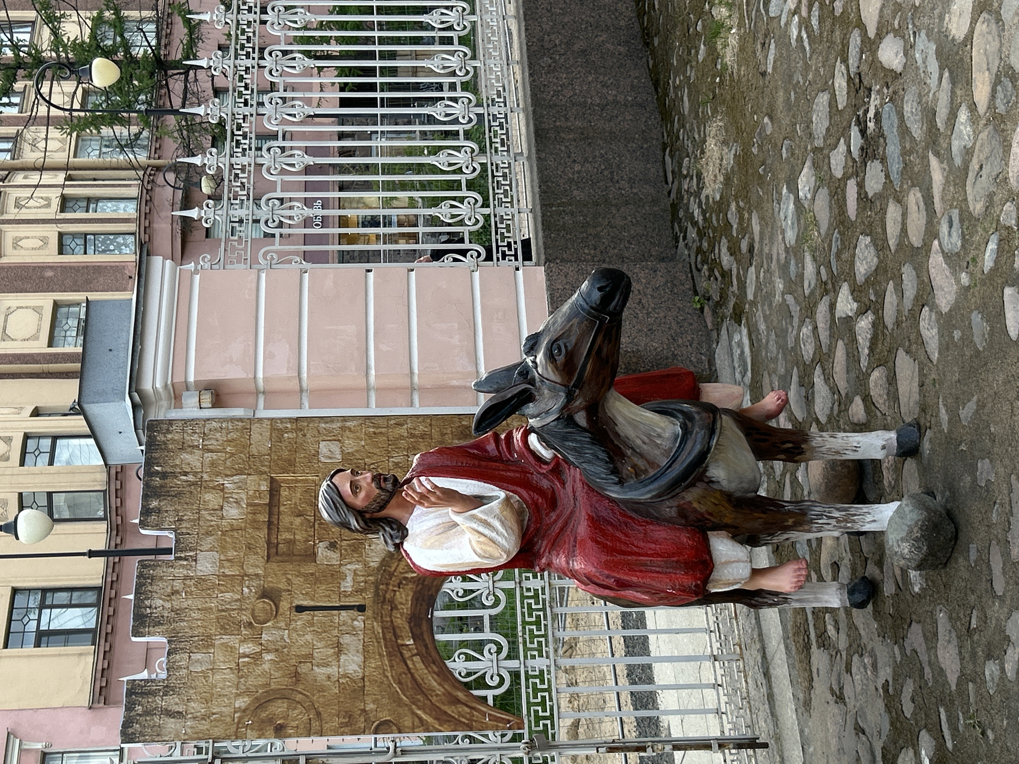 A statue of Jesus riding a donkey stands in front of a building in Saint Petersburg, Russia. The statue is brightly colored, with Jesus wearing a red robe and the donkey a brown and tan hue. The building behind the statue is beige and pink with multi-paned windows. A decorative white fence runs along the front of the building and in front of the statue. The ground is paved with cobblestones.