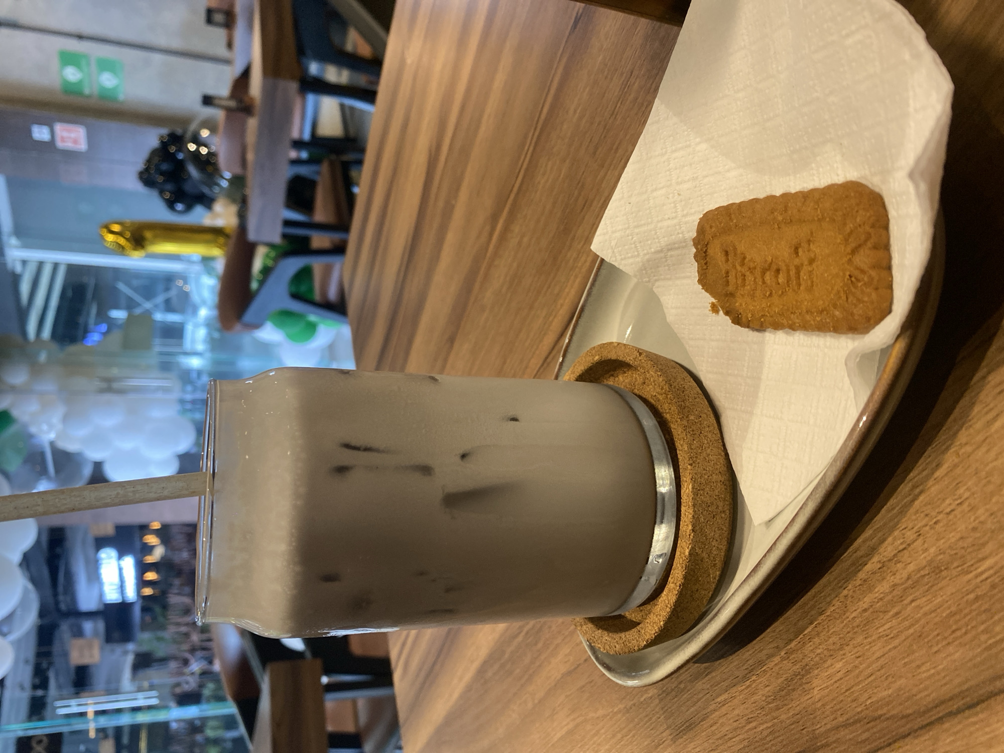 A close-up, canted indoor shot shows a rectangular glass of light brown iced coffee with a straw, sitting on a round cork coaster on a small white plate, with a rectangular cookie wrapped in a white napkin on the plate, on a wooden table.
The coffee glass is mostly clear and has visible ice cubes, and the straw is a light brown color. The cork coaster is circular. The white plate has a slightly raised edge and appears to be glazed. The rectangular cookie has a brown color with text imprinted in the middle.
The wooden table is light brown and appears to have a veneer or laminate finish. In the blurred background, there are hints of an urban environment, including people, balloons (white and gold), and architectural details suggesting a city setting, but no identifying markers are visible. The exact location and city are not discernible from this image.