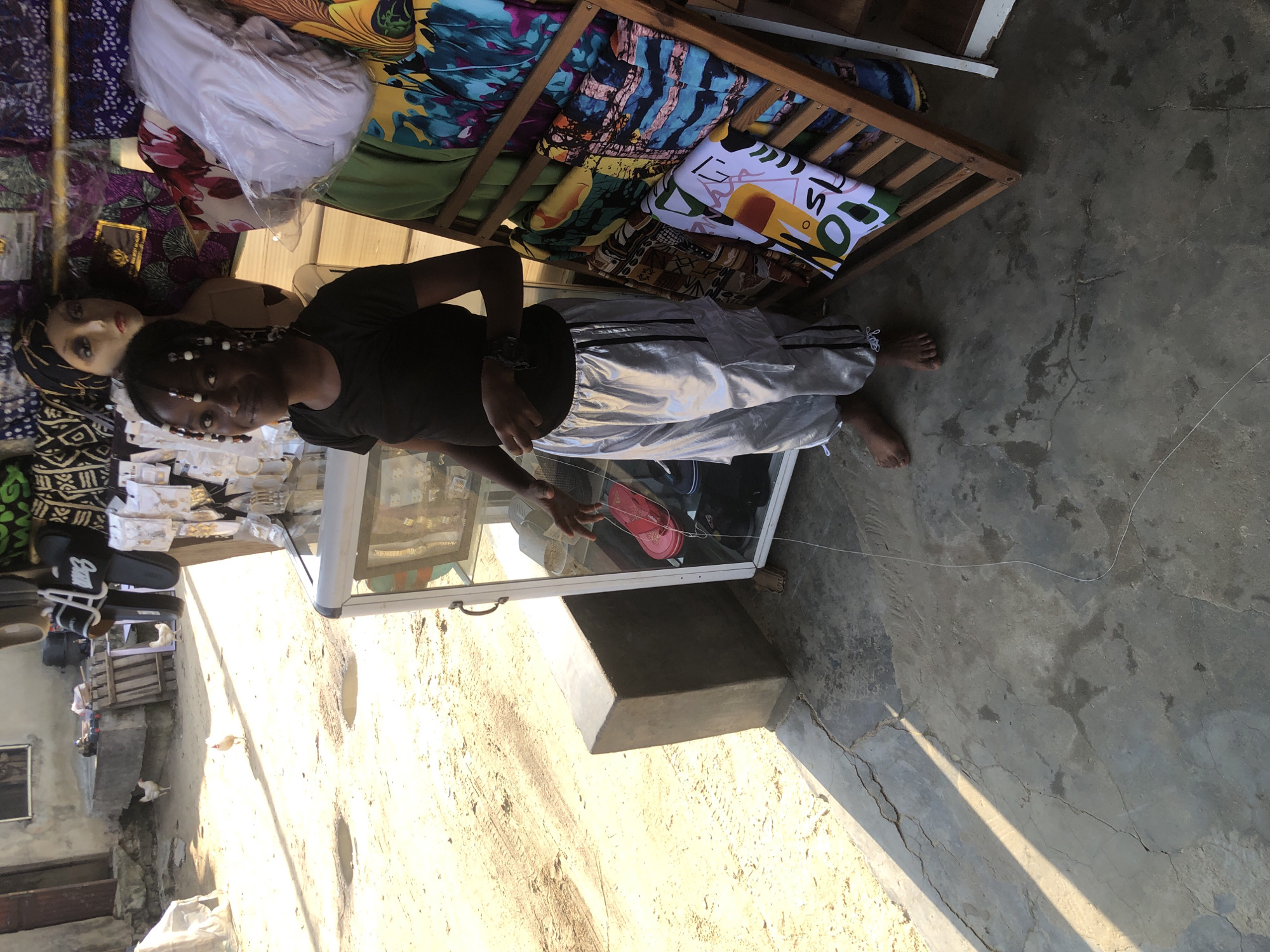 A young Black girl, with beads in her hair, stands barefoot on a cracked concrete floor in a market stall. She is wearing a black t-shirt and shiny silver pants. To her left, a glass display case contains red and blue flip-flops, while a wooden rack to her right is filled with brightly patterned fabrics. In the background, several chickens peck at the dusty ground outside a weathered building. The scene appears to be an outdoor market in Ado, Nigeria, characterized by its warm, natural lighting and the presence of local goods and livestock.