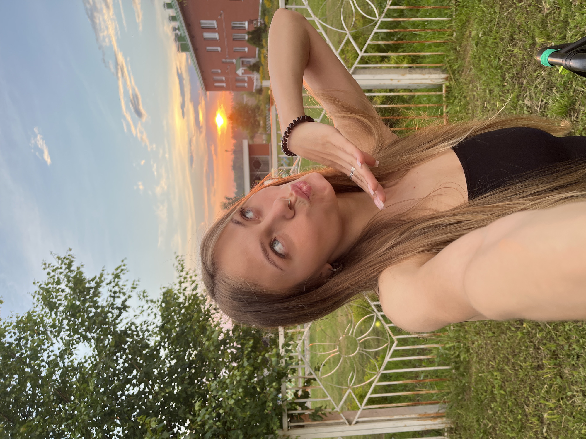 A young Caucasian woman with long blonde hair is taking a selfie outdoors at sunset. She is wearing a black tank top and has her hair pulled back over her left shoulder. Her left arm is extended to her side, and her right hand is held up to her face, obscuring part of her mouth. Her lips are puckered, and she is looking up and to the right. In the background, a white metal fence runs horizontally across the image. Beyond the fence, there is a patch of green grass, a tree on the left, and a red brick building with a green roof on the right. The sky is a mix of blue and orange, with the sun setting in the distance. This scene is taking place in the city of Fokino, Russia.