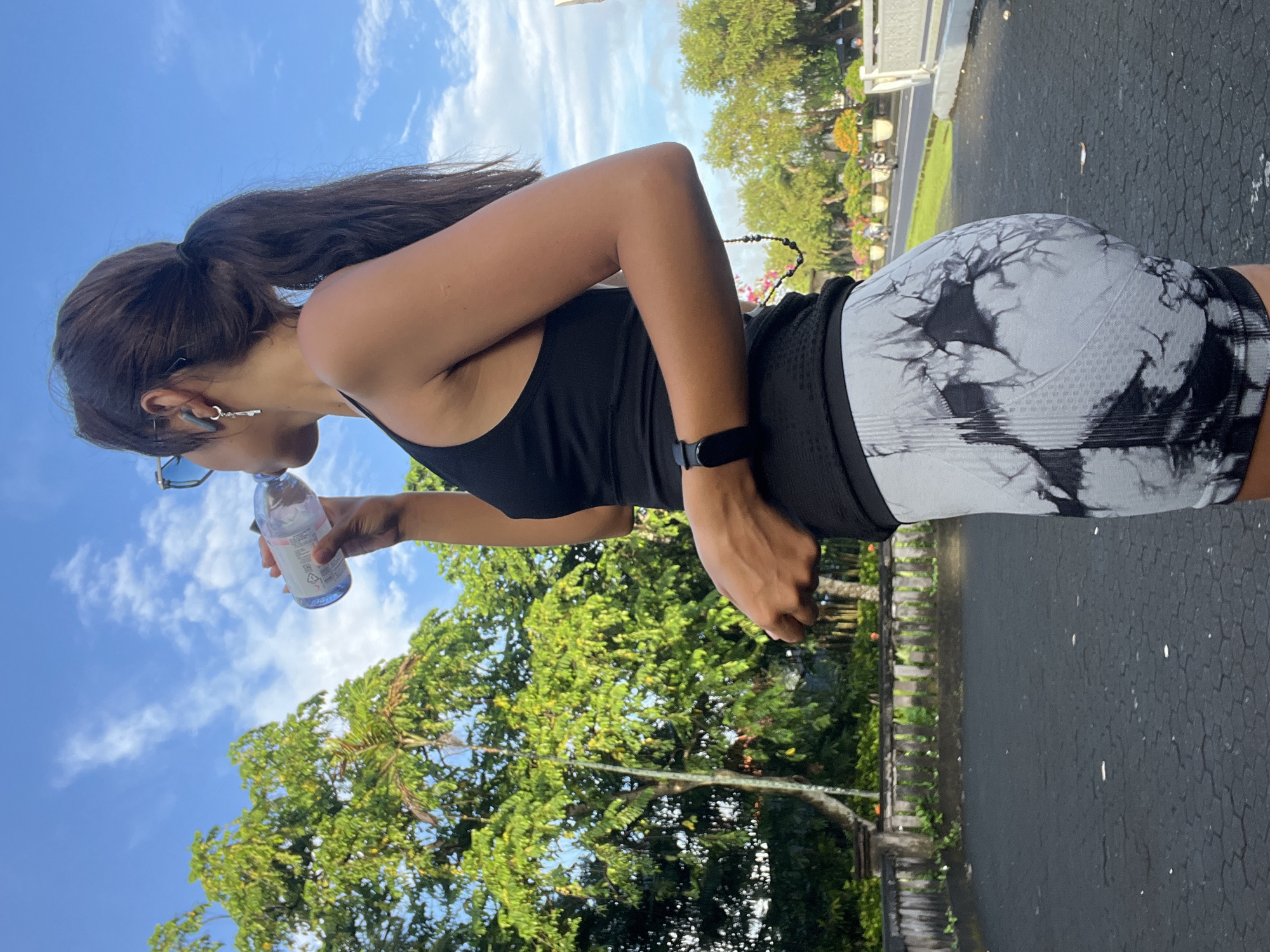 An outdoor, eye-level shot captures a person, likely female, in a side profile, drinking from a clear plastic water bottle. She has dark hair styled in a high ponytail and is wearing a black athletic tank top and black and white tie-dye patterned cycling shorts or leggings. A black smartwatch is visible on her left wrist, and sunglasses hang from her neck.
The background features a vibrant blue sky with scattered white clouds, indicating a clear, sunny day. Lush green tropical trees are prominent, with some closer to the foreground and others forming a dense canopy further back. A paved surface, possibly a road or path with hexagonal patterns, is visible to the right. A low stone fence or wall with additional greenery runs parallel to the paved area in the mid-ground.
The person is actively rehydrating, holding the bottle to her mouth. The word "AQUA" is faintly visible on the water bottle. The scene exudes a sense of outdoor activity in a warm, pleasant climate, consistent with the location of Kuta Selatan, Indonesia.
