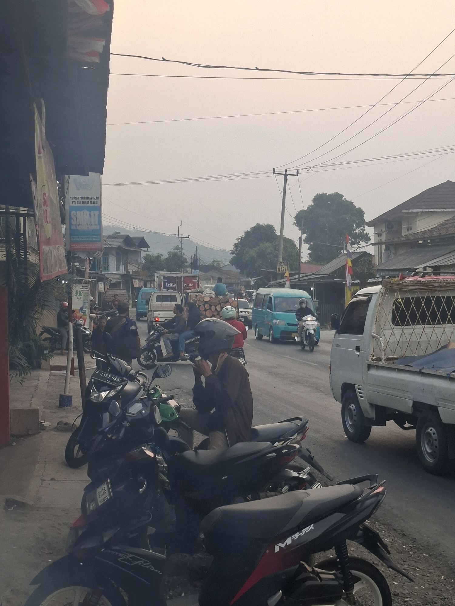 The image depicts a street scene in Cijalingan, Indonesia, characterized by a hazy sky and a mix of vehicles and pedestrians. The foreground features several parked motorcycles, with one black motorcycle prominently displaying a license plate that reads "F 2152 DAA". To the right, a white pickup truck is visible, its cargo bed containing a blue tarp and a diamond-patterned metal frame.
In the background, a bright blue minivan is in motion, driven by a person wearing a helmet. Other vehicles, including a white pickup truck and another light blue vehicle, are also present on the road. A group of people are gathered on the left side of the street, some standing near buildings and others interacting near the motorcycles. A banner for "YAYASAN PENDIDIKAN ISLAM" and "SMK IT" is displayed on a building. The environment includes power lines overhead, trees, and distant hills. The overall atmosphere suggests a busy, everyday street in a semi-urban or rural setting.