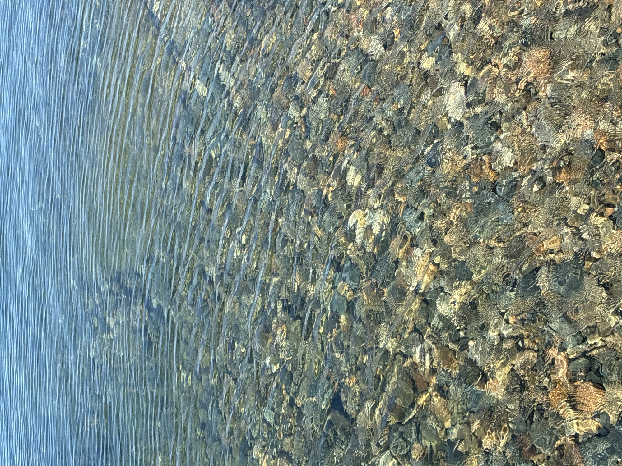 The image displays a close-up, overhead view of the surface of a body of water, likely Lake Baikal, near Listvyanka, Russia. The water is clear and shallow, revealing a bottom covered with numerous smooth, rounded stones of various sizes and colors, including shades of grey, brown, and beige. Gentle ripples disturb the water's surface, creating distorted reflections of the stones below and a shimmering effect. The upper portion of the image transitions to a deeper blue expanse of water with more pronounced, parallel ripples. No people, animals, or man-made objects are visible in the frame. The scene is natural and tranquil, focusing on the texture and clarity of the water and the submerged stones.