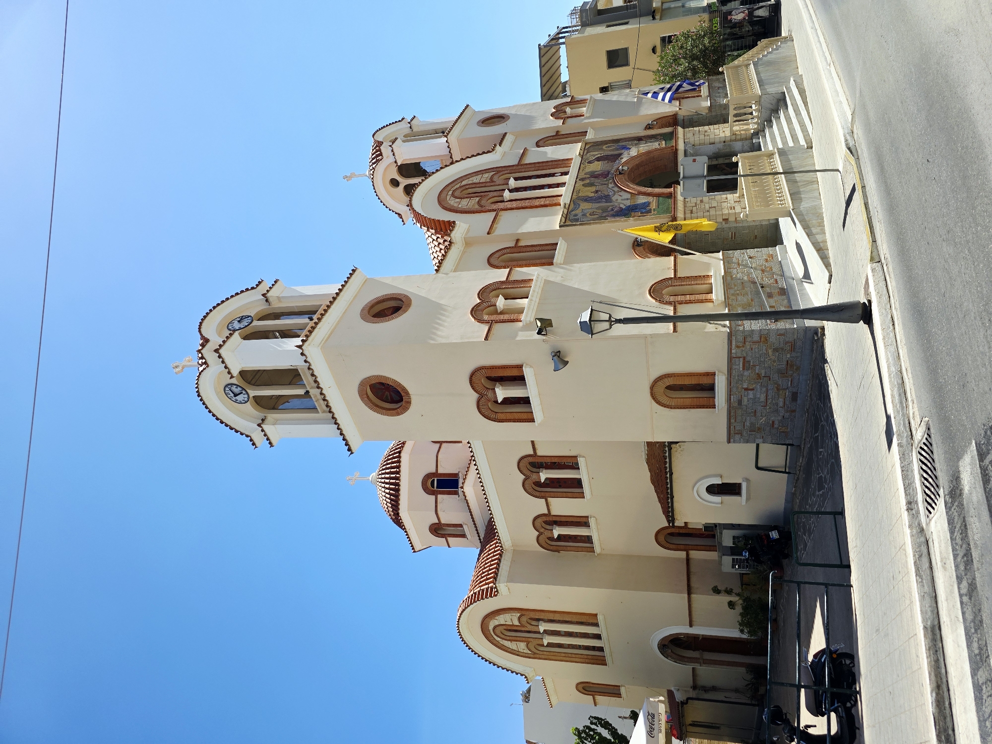 This is a medium-angle, eye-level, exterior shot of a church, likely a Greek Orthodox church, on a clear, sunny day in Agios Nikolaos Municipal Unit, Greece. The church has a cream-colored facade, red tile roofs, and numerous arched windows. The main entrance is flanked by stairs and has a mosaic above it. A tall bell tower with a clock is visible. Two flags, one yellow and one blue and white, are displayed in front of the church. A lamppost stands on the sidewalk in front of the building. To the left, a white umbrella with a Coca-Cola logo is visible, suggesting a nearby cafe or shop. A black scooter is parked on the left side of the image, partially obscured by a fence. The street in front of the church is paved, with a sidewalk and a drain visible. The sky is a bright, clear blue.