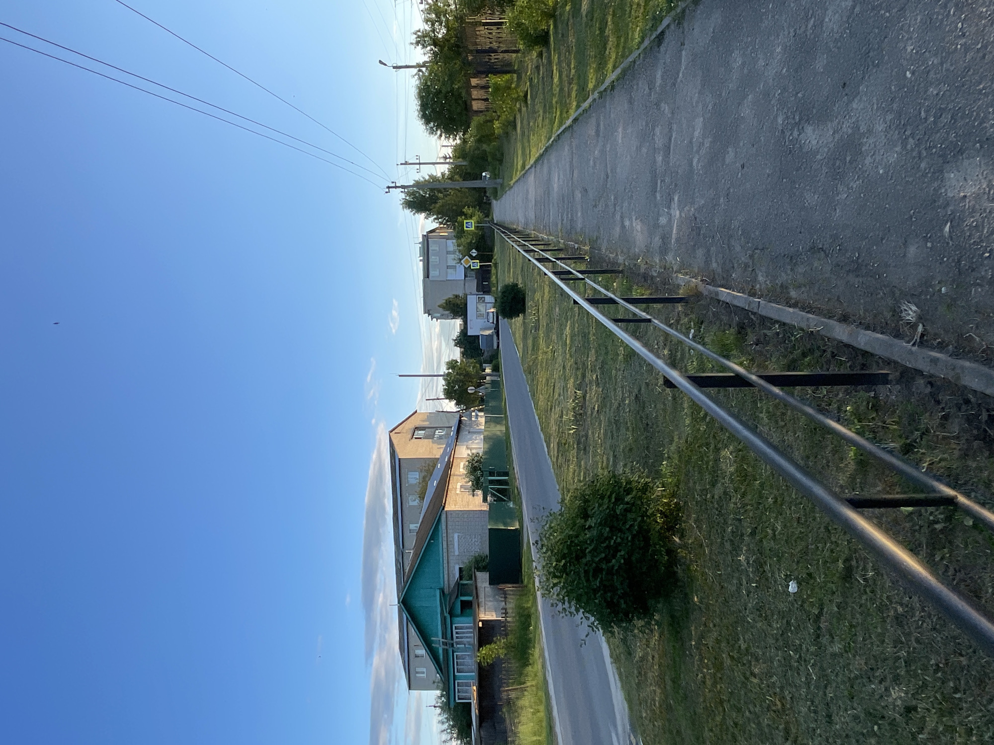 The image depicts a street scene in the settlement of Krasnye Baki, Russia, under a clear blue sky with scattered clouds. The perspective is from a paved sidewalk bordered by a black metal railing on the left. To the left of the sidewalk is a grassy area with a few bushes, and beyond that, an asphalt road. Along the road, on the left side, are several buildings. The closest building is a single-story structure with a green roof and lighter-colored brick walls, appearing to be a residential house. Behind it, a taller, multi-story building with a flat roof is visible. On the right side of the sidewalk, there is a continuous line of trees and a dark metal fence. Electrical wires and poles are present along the right side of the street. Two yellow pedestrian crossing signs are visible in the distance on the right side of the road. The environment suggests a quiet, possibly rural or suburban area. There are no people visible in the image.