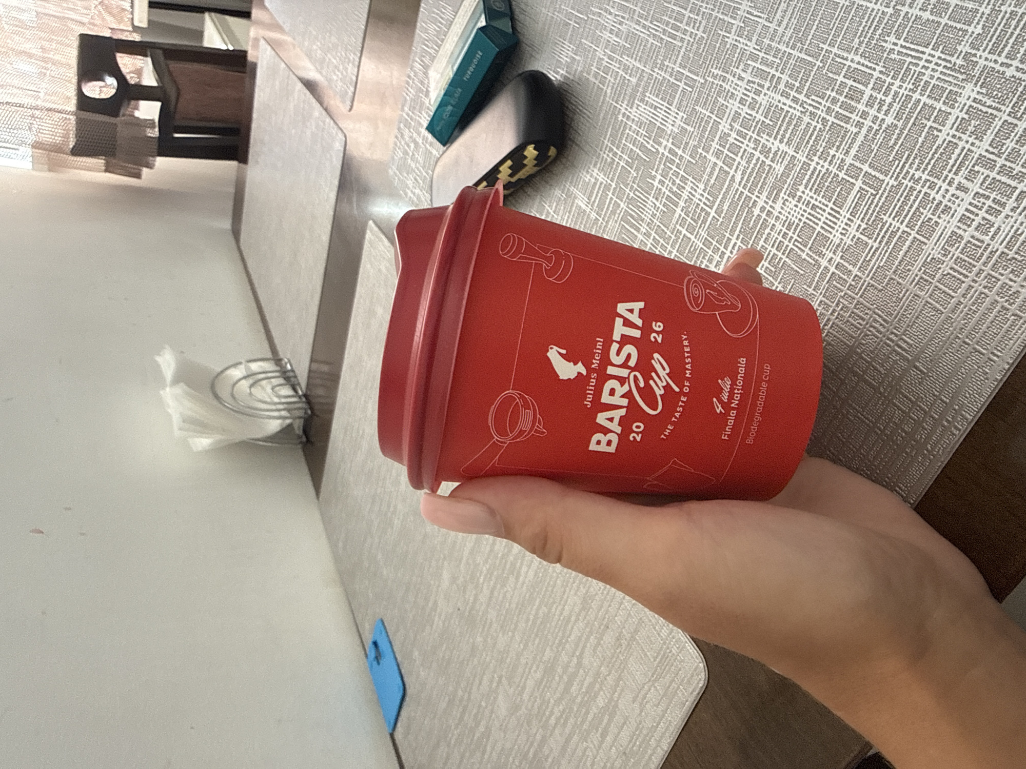 The image depicts a close-up of a person's hand holding a red disposable coffee cup with "BARISTA CUP" printed on it. The cup has the number "20" and "26" along with the text "THE TASTE OF MASTERY". Below this text, it says "4 iulie Finala Națională" and "Biodegradable cup". The background shows a table with a textured placemat, a blue object on the table, a napkin holder with napkins, and a portion of a wooden chair and table in the distance. The scene appears to be indoors, possibly in a cafe or restaurant. There is no discernible information to identify the specific location within Chișinău, Moldova.