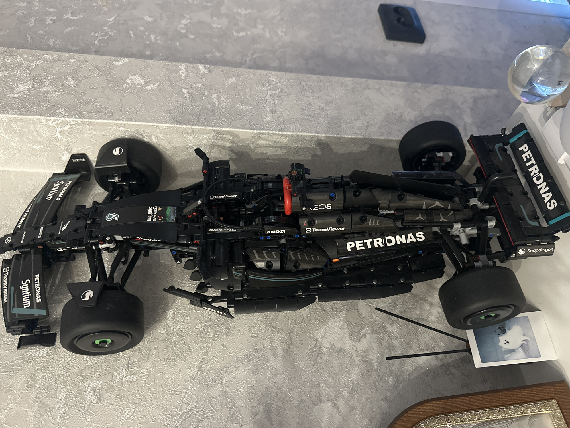 A detailed, black model of a Formula 1 racing car, resembling a Mercedes-AMG Petronas F1 W12, dominates the frame. The model, likely constructed from LEGO Technic elements, is highly intricate, showcasing an open-wheel design, suspension components, a detailed engine block, and various aerodynamic wings and diffusers. It is positioned on a textured, light grey surface, possibly a wall or shelf, and the image itself is rotated 90 degrees clockwise.
Prominent branding visible on the car includes "PETRONAS," "Syntium," "TeamViewer," "INEOS," "AMD," and "Snapdragon," along with associated logos such as the Mercedes star and a stylized 'S' on the wheel covers. The car features light blue accents on its livery.
In the upper right portion of the image (due to rotation), a clear glass sphere reflects its surroundings. Further up, a dark rectangular object is mounted on the wall. In the lower right, beneath the car's rear wheel, a small instant photograph (Polaroid-style) featuring a white animal, possibly a cat or dog, is propped up by two thin black rods. Part of a decorative wooden frame with silver embellishments is visible in the very bottom of the shot. The setting appears to be indoors, under artificial lighting. No activity is taking place; the image captures a static display.