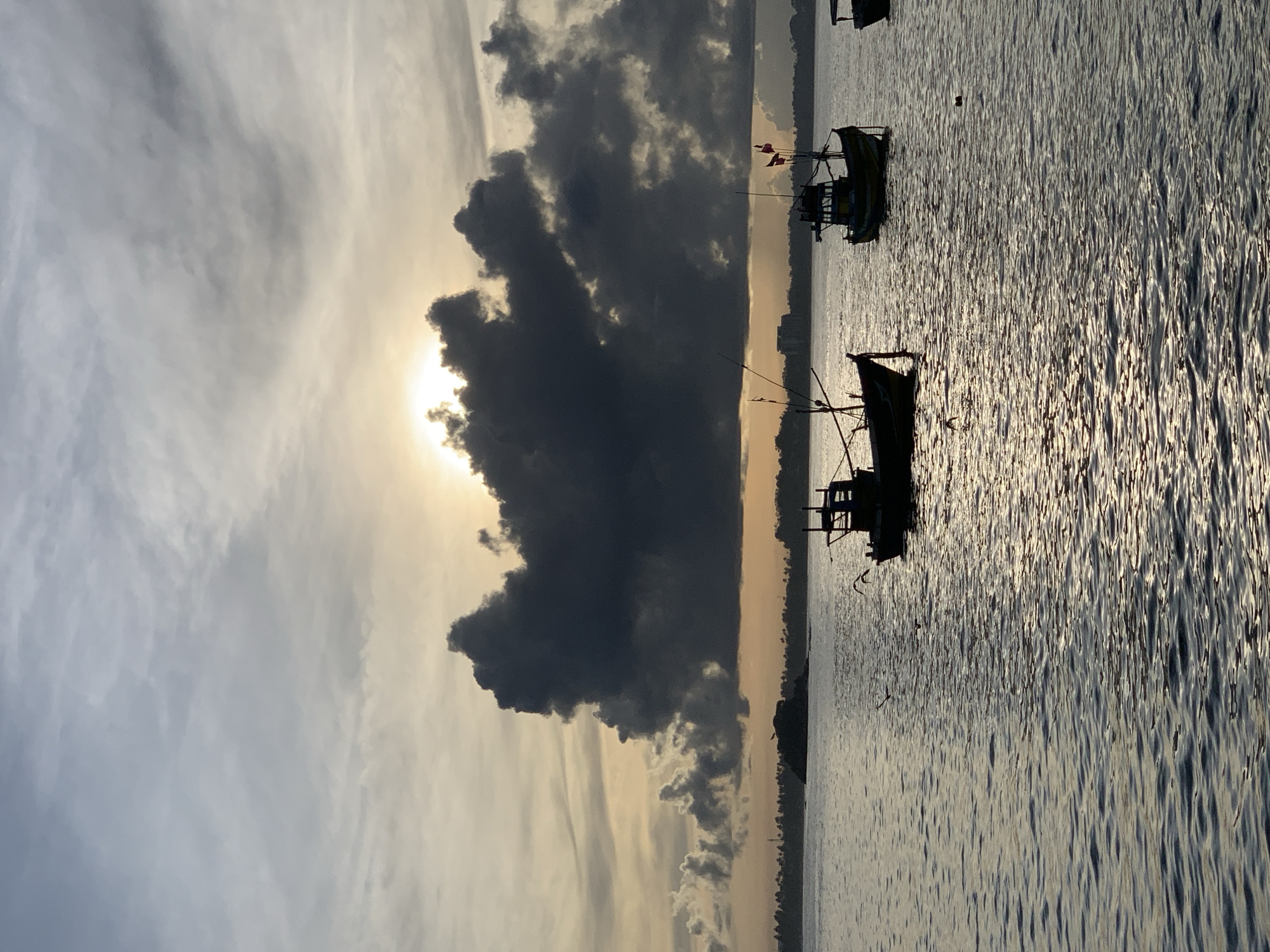 The image depicts a serene coastal scene at either sunrise or sunset in Weligama, Sri Lanka. The sun, a brilliant source of light, is positioned low in the sky on the left side of the frame, partially obscured by a large, dramatic dark cloud formation. This creates a striking silhouette effect, casting the cloud as a prominent dark mass against the bright sky. The sky transitions from a warm, golden glow near the sun to softer grey and white wispy clouds extending towards the upper right.
On the right side of the frame, two traditional fishing boats are silhouetted against the shimmering water. Their distinctive structures, including masts, rigging, and parts of their cabins, are visible as dark shapes, enhancing the dramatic contrast of the low light. The water surface is textured with numerous small ripples, catching the golden light and creating a sparkling, wavy pattern that extends across the right half and foreground of the image. A faint, distant coastline or landmass can be seen along the horizon line, which runs horizontally across the mid-section.
The boats appear stationary on the calm water, suggesting a moment of tranquility before or after active fishing. The weather is partly cloudy, with the dark cloud dominating the central-left sky. The overall mood is peaceful and contemplative, characteristic of dawn or dusk over a body of water. There is no visible text in the image.