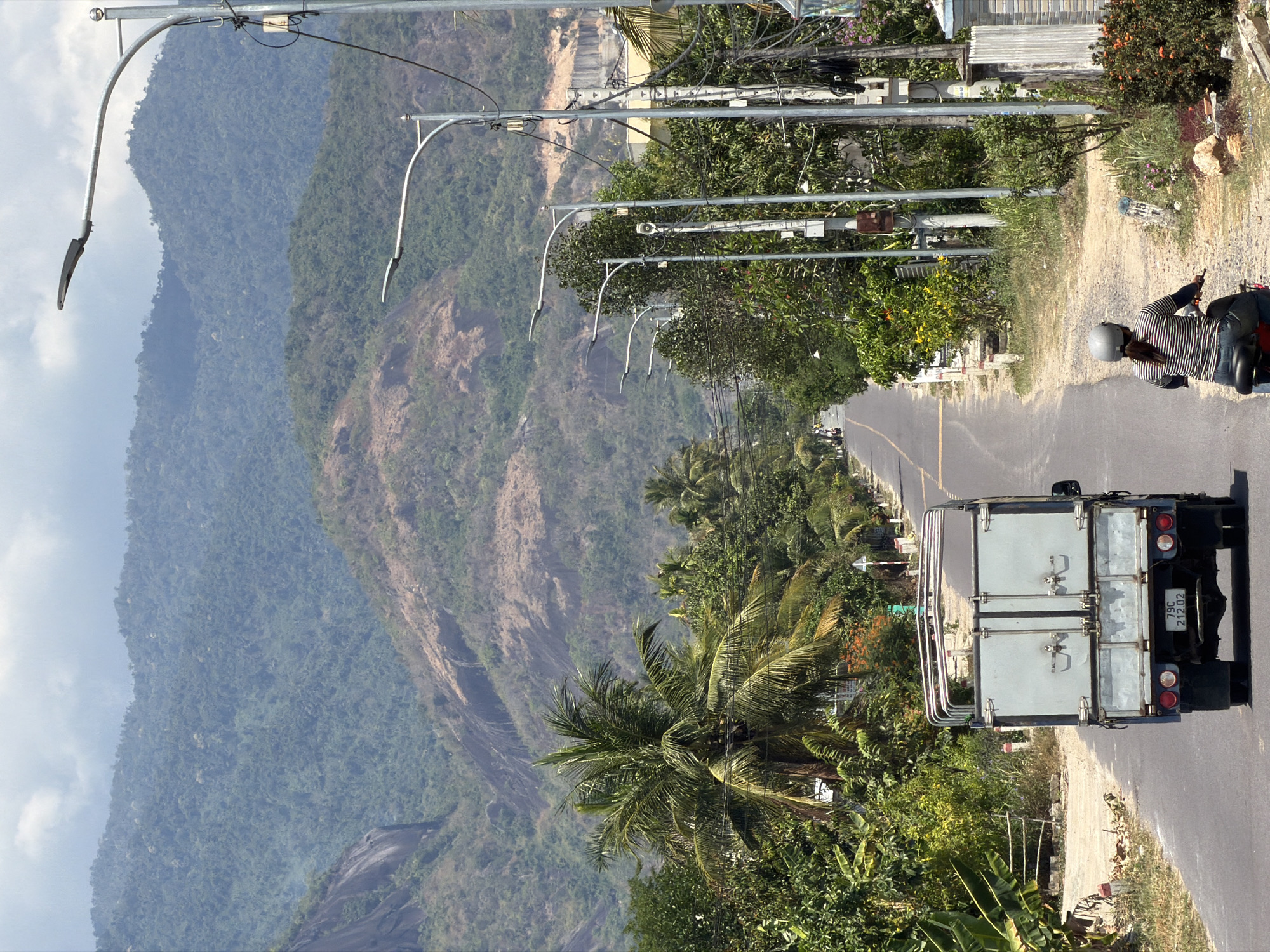 A vertically oriented daytime shot in Diên Thọ, Vietnam, shows a road winding past a mountain. On the right side of the road, a white truck with visible license plate "79C-212.02" is parked. A person on a motorcycle is nearby. Power lines and streetlights run along the side of the road. Trees and other foliage line the roadside. The landscape slopes upward to the left, where a mountain covered in trees and vegetation is visible. The sky is visible in the top left corner.