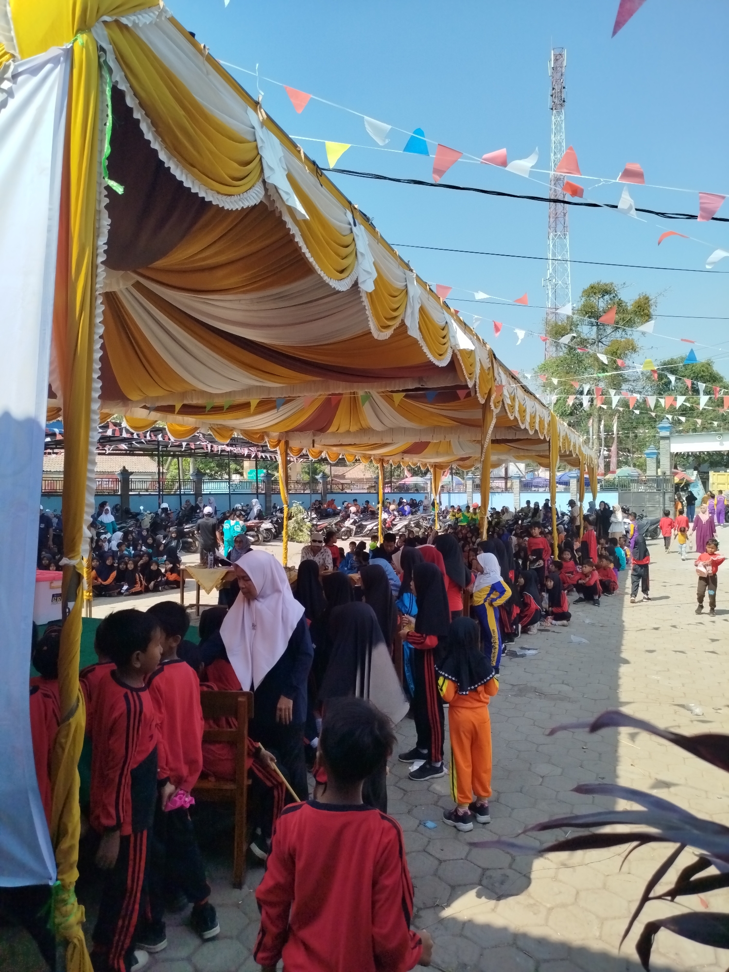 This is a medium shot, eye-level, outdoor photograph taken in a sunny, open-air setting, likely in Parseh, Indonesia. The image depicts a gathering of people, predominantly children and some adults, under a large, yellow and white tent. The tent has a scalloped edge and is supported by poles, casting shadows on the ground.
The foreground is occupied by several children wearing red uniforms with black stripes. One child in the very front is facing away from the camera, showing the back of their red uniform. To their left, another child in the same uniform is looking to the side. A girl in an orange tracksuit is standing on the right side of the frame.
Behind these children, the crowd continues under the tent and extends into the background. Many individuals are seated or standing. Some adults are present, including a woman wearing a light pink headscarf. There are motorcycles parked in the background, suggesting a public event or gathering.
The environment is paved with interlocking bricks. In the background, a tall telecommunications tower is visible against a clear blue sky. Small, colorful triangular flags are strung across the sky and between poles, indicating a festive occasion. Trees and buildings are also visible in the distance. The overall atmosphere appears to be that of a community event or school activity.