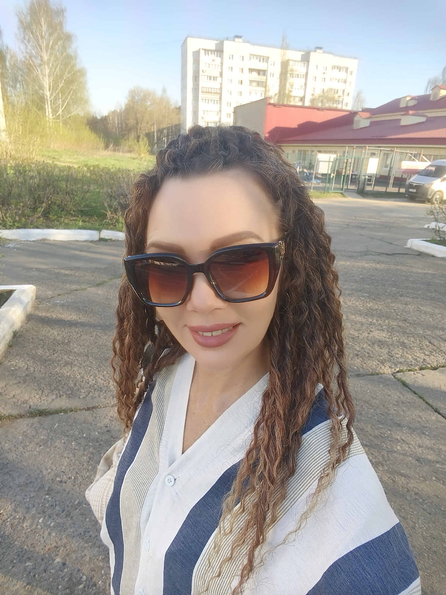 A woman with curly brown hair and sunglasses stands on a paved surface in an urban setting. She is wearing a striped shirt with blue and white alternating stripes. Behind her, there is a green area with trees and a building with a red roof. In the background, a taller white apartment building is visible under a blue sky. A car is parked near the building with the red roof. The location appears to be in an area with both residential buildings and green spaces.