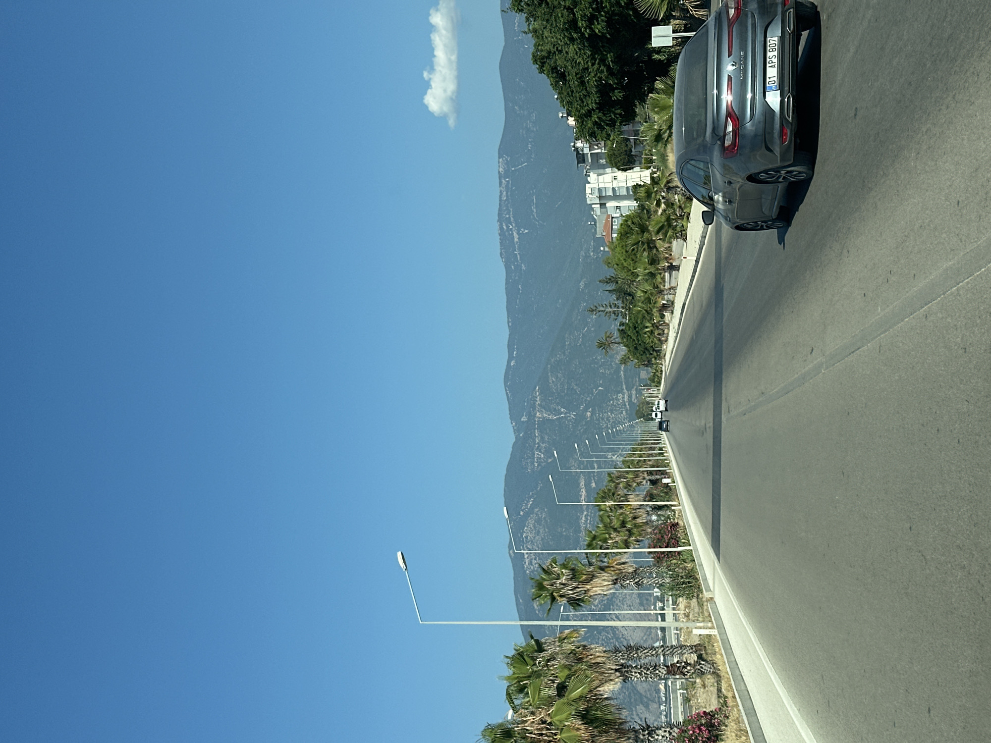 A car, a gray Renault Megane with license plate "01 APS 807", is driving on a two-lane road in Finike, Turkey. The road is lined with palm trees and streetlights on the left side. In the background, there are mountains and some buildings. The sky is clear and blue with a single white cloud. The image appears to be taken from another vehicle on the road.