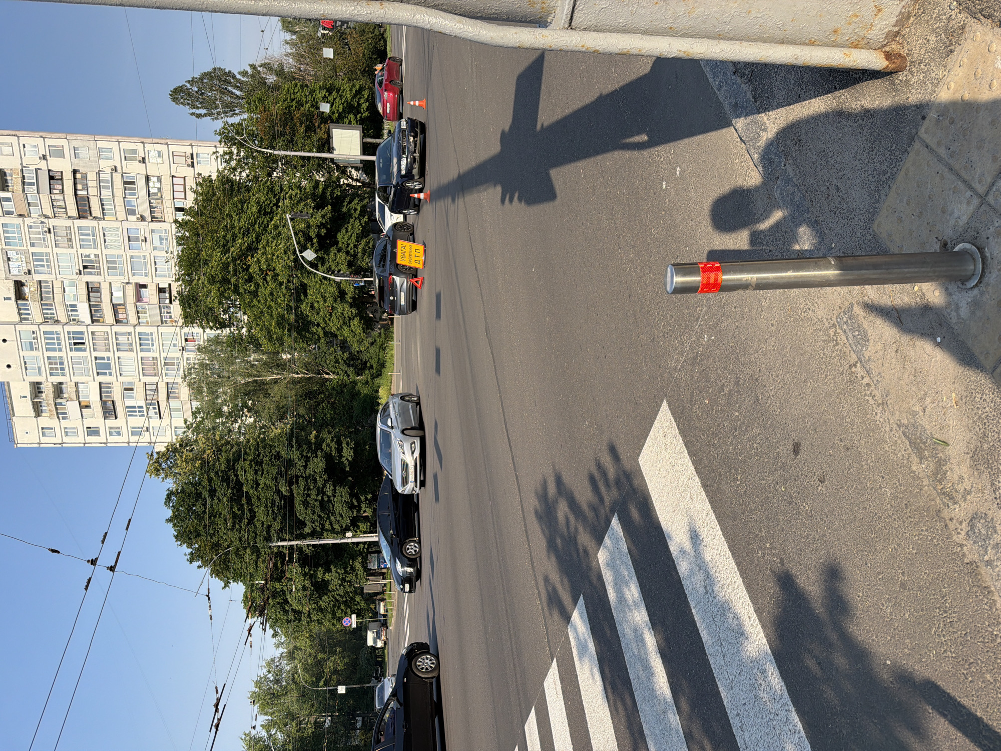 A vertical, low-angle shot shows a street scene in Kyiv, Ukraine. The foreground features a pedestrian crossing with bold white stripes. To the right of the crossing is a silver metal pole with red reflective tape, set into a concrete sidewalk. Shadows of trees and what appear to be people are cast across the road and sidewalk, suggesting a sunny day.
Several cars are visible on the road, including a black SUV and a black sedan stopped at the crossing, a white SUV, and a red car further down the street. One black car has a yellow warning sign with red lettering that reads "УВАГА! ОБЕРЕЖНО ДТП" (Attention! Caution! Accident). Orange traffic cones are placed near some of the vehicles, indicating a possible obstruction or hazard.
In the background, a large, multi-story apartment building with numerous balconies and windows dominates the left side of the frame. Dense green trees line the right side of the street, creating a stark contrast between the urban architecture and the natural elements. Overhead, a complex network of electrical wires, likely for trolleybuses or trams, crisscrosses the sky. The sky is a clear, pale blue, indicating fair weather. The lighting suggests it is daytime, possibly late afternoon due to the long shadows.