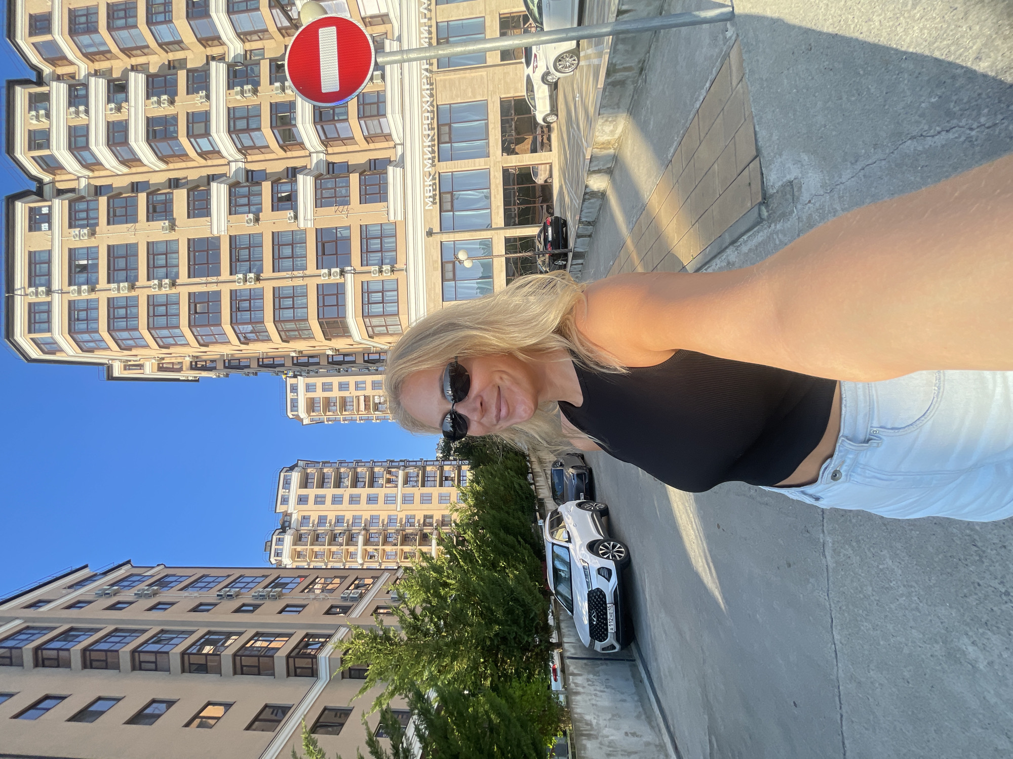 A blonde woman, wearing sunglasses, a black tank top, and light blue jeans, captures a selfie outdoors under a clear blue sky. She is positioned on a paved surface, extending her right arm towards the camera with a slight smile. To her left, a low concrete wall fronts a row of manicured evergreen shrubs, behind which stand multi-story, beige apartment buildings with dark-framed windows and balconies. A white Chery Tiggo SUV with Russian license plate "A 112 HE 45" is parked by the wall, followed by a dark sedan.
On the right side of the image, another series of modern, multi-story buildings with similar beige facades and numerous exterior air conditioning units line a street. A prominent red "No Entry" traffic sign on a pole is visible in the foreground on the right. Behind the sign, one building features a ground-level commercial facade with the Cyrillic inscription "МВК МИКРОХИРУРГИИ ГЛАЗА" (MVK Microsurgery of the Eye) above its large windows. Sunlight casts defined shadows, indicating bright daytime conditions. The architecture and identified clinic point to the location as Ulitsa Gagarina, 78, Tsentral'nyy rayon, Sochi, Krasnodarskiy Kray, Russia.