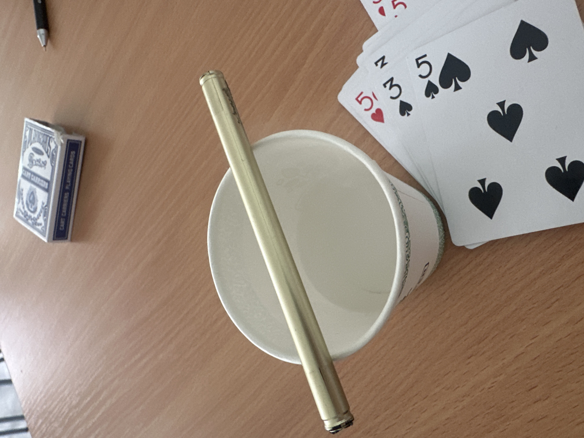 The image captures a top-down view of several items arranged on a light brown wooden surface, possibly a desk or table. In the upper left corner, a black pen with a metallic tip is partially visible. Below it and slightly to the right, a blue deck of playing cards in its box is positioned. The box features ornate white designs, and the word "BICYCLE" is visible on its front face. On the side of the box, text reads "CART CARRIERS PLAYING CARDS".
Dominating the center of the frame is a white paper cup, oriented with its opening facing upwards and slightly towards the viewer. A golden-colored, cylindrical object, resembling a pen or a long metal tube, rests horizontally across the rim of the cup. The object has a metallic sheen.
In the upper right portion of the image, a fan of playing cards is spread out. The visible cards include:
- A card showing a red "5", likely a 5 of Hearts.
- A card displaying an upside-down "N" and a "3" next to a black spade symbol, indicating a 3 of Spades.
- A card clearly showing a black "5" next to a black spade symbol, identifying it as a 5 of Spades.
- Other cards further down reveal additional black spade symbols, suggesting more spade cards in the hand.
The overall scene suggests a casual setting, perhaps during a game or a break. The lighting indicates it is likely daytime or a well-lit indoor environment. No people are visible, and no specific emotions are conveyed by the inanimate objects.