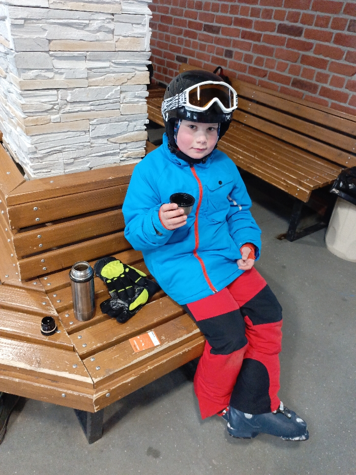 In the city of Svetloe, Russia, a young child wearing ski attire is seated on a wooden bench. The child is a male, dressed in a black ski helmet with white goggles pushed onto it, a blue ski jacket with a red zipper, red and black ski pants, and blue ski boots. He holds a small, dark-colored cup in his right hand, looking directly forward. To his left, on the same bench, are a silver thermos flask with its black cap placed beside it, a pair of black and yellow ski gloves, and an orange rectangular pass or ticket. Behind the child and to his left is a pillar or wall section covered in light-colored stone veneer. To his right and further back is a reddish-brown brick wall with a dark wooden bench positioned against it. The floor is a grey, possibly concrete, surface. The scene suggests a rest period in an indoor or sheltered area of a ski resort or facility.