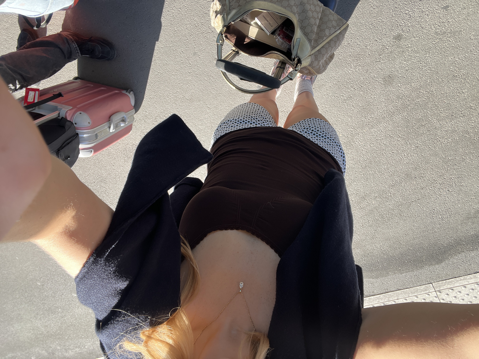 A high-angle shot shows a person standing on a paved surface, possibly a sidewalk or street, in an urban setting. The subject is wearing a brown top and patterned shorts, partially covered by a dark blue cardigan. A gold necklace with a pendant is visible around the neck. To the left, a pink suitcase is positioned next to a person wearing dark pants and black shoes. A Gucci handbag lies on the ground at the top of the frame. The items inside the bag seem to include cigarettes. The location might be inferred as Paris, France.