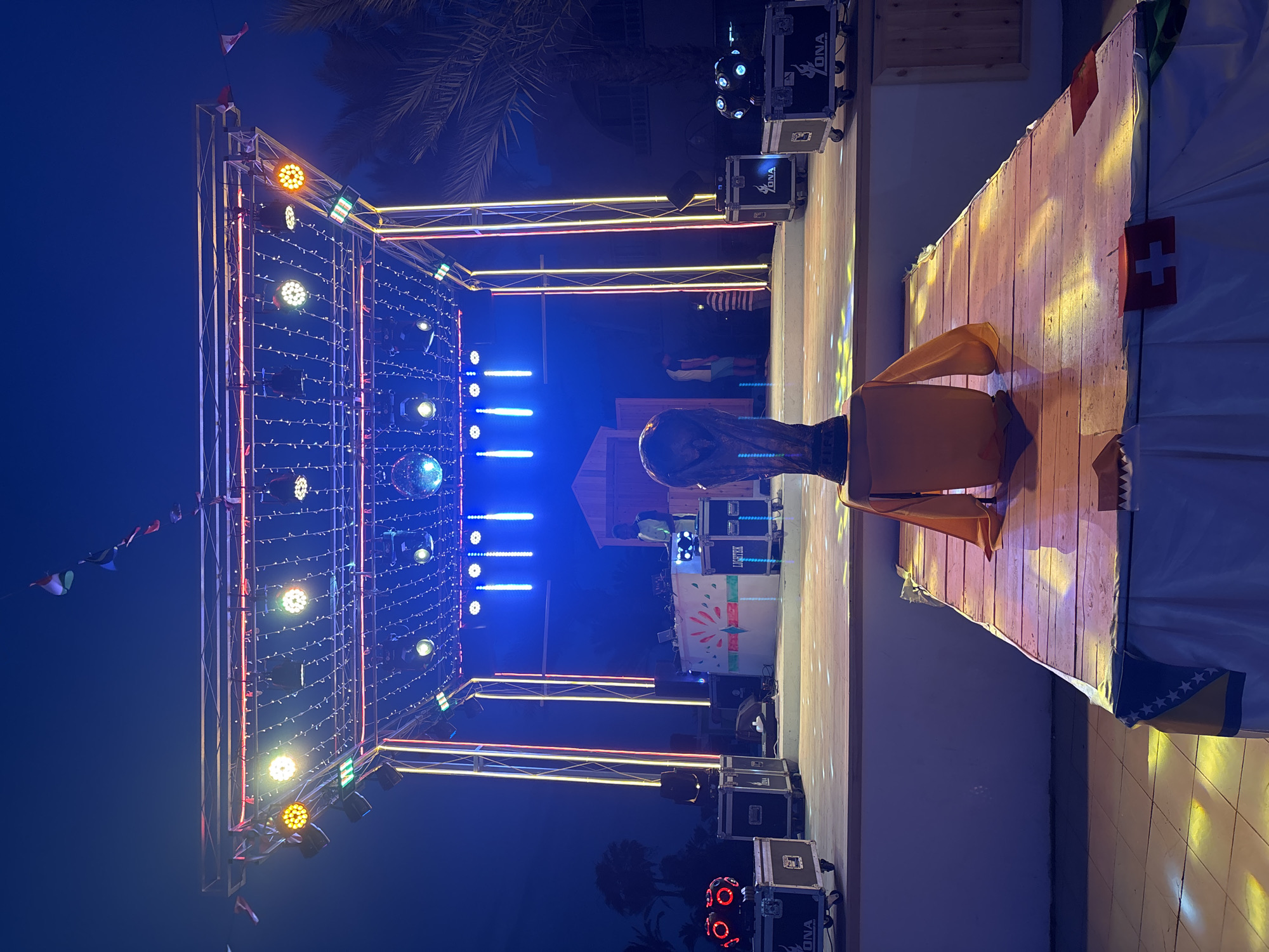 An outdoor stage setup is illuminated with colorful lights and string lights at dusk or night in Taguermess, Tunisia. A DJ booth with a DJ and two people behind it is positioned on the elevated stage. Several speakers and lighting equipment are visible on the stage and in front of it. A large trophy resembling the FIFA World Cup is prominently displayed on a wooden platform in the foreground. Flags of various countries are draped around the platform and hanging above the stage. Palm trees and buildings are faintly visible in the background.