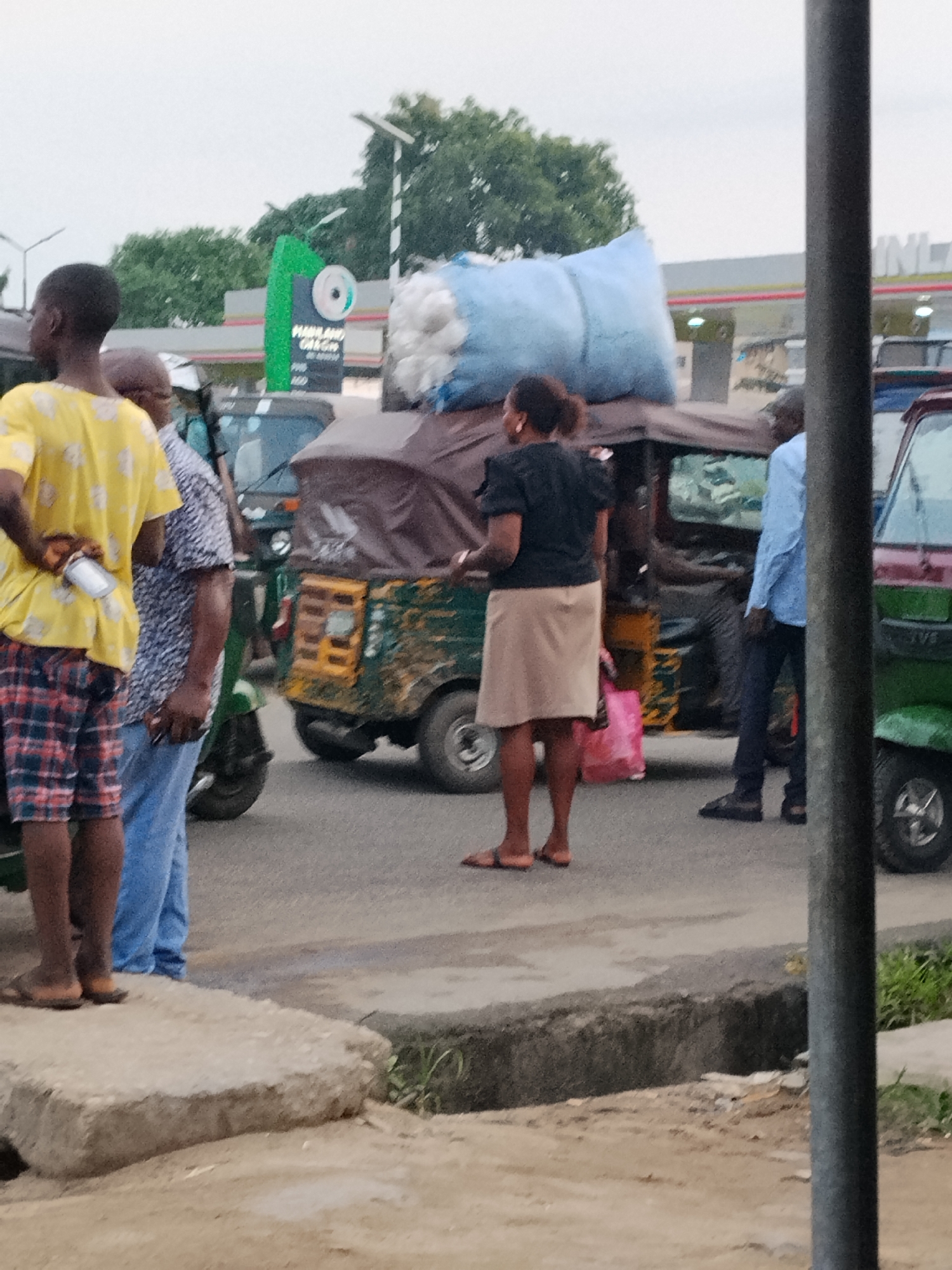 The image depicts a street scene in what appears to be Aba, Nigeria. Several individuals are present, including a person in a yellow shirt and plaid shorts holding a silver object, a person in a patterned shirt and jeans, a woman with a tan skirt and dark top carrying a pink bag, and a man in a blue shirt. These people are in proximity to motorized tricycle vehicles (tuk-tuks), with one heavily loaded with a large white sack. A green gas station sign with the words "HAMLAND GAS" is visible in the background. The environment suggests a busy street with visible signs of urban life.