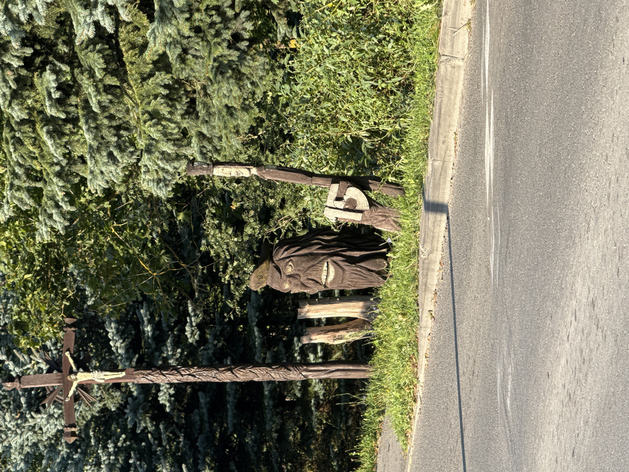 In the city of Galinė, Lithuania, a wooden crucifix with a figure of Jesus Christ stands tall on the left side of the frame, set against a backdrop of dense green trees. To the right of the crucifix, a wooden carving of a bearded figure with an open mouth and large eyes is positioned. Further to the right, another wooden post is visible with a carved face at the top and the number "15" with a spider motif at the bottom. The foreground shows a paved road with a concrete curb and a strip of green grass. The scene appears to be an outdoor setting, possibly near a roadside or in a rural area.