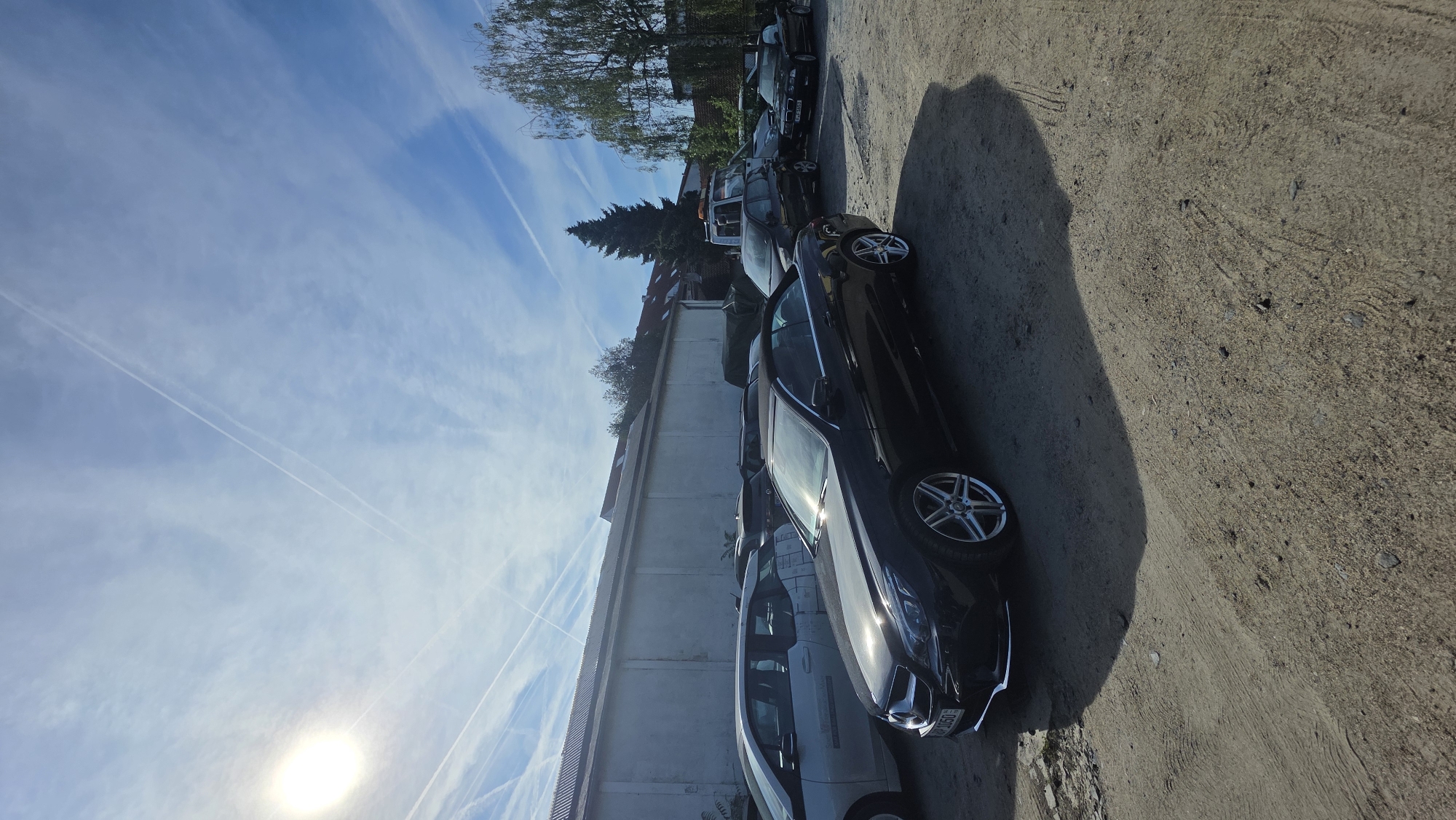 The image shows a black convertible Mercedes-Benz parked on a dirt lot. The car is in the foreground, with other vehicles parked behind it, including a white hatchback and a tow truck. The lot appears to be a storage area or a used car dealership. In the background, there is a long, white building with a metal roof, and some trees. The sky is blue with scattered clouds and contrails from airplanes. The sun is visible in the upper left corner of the image.