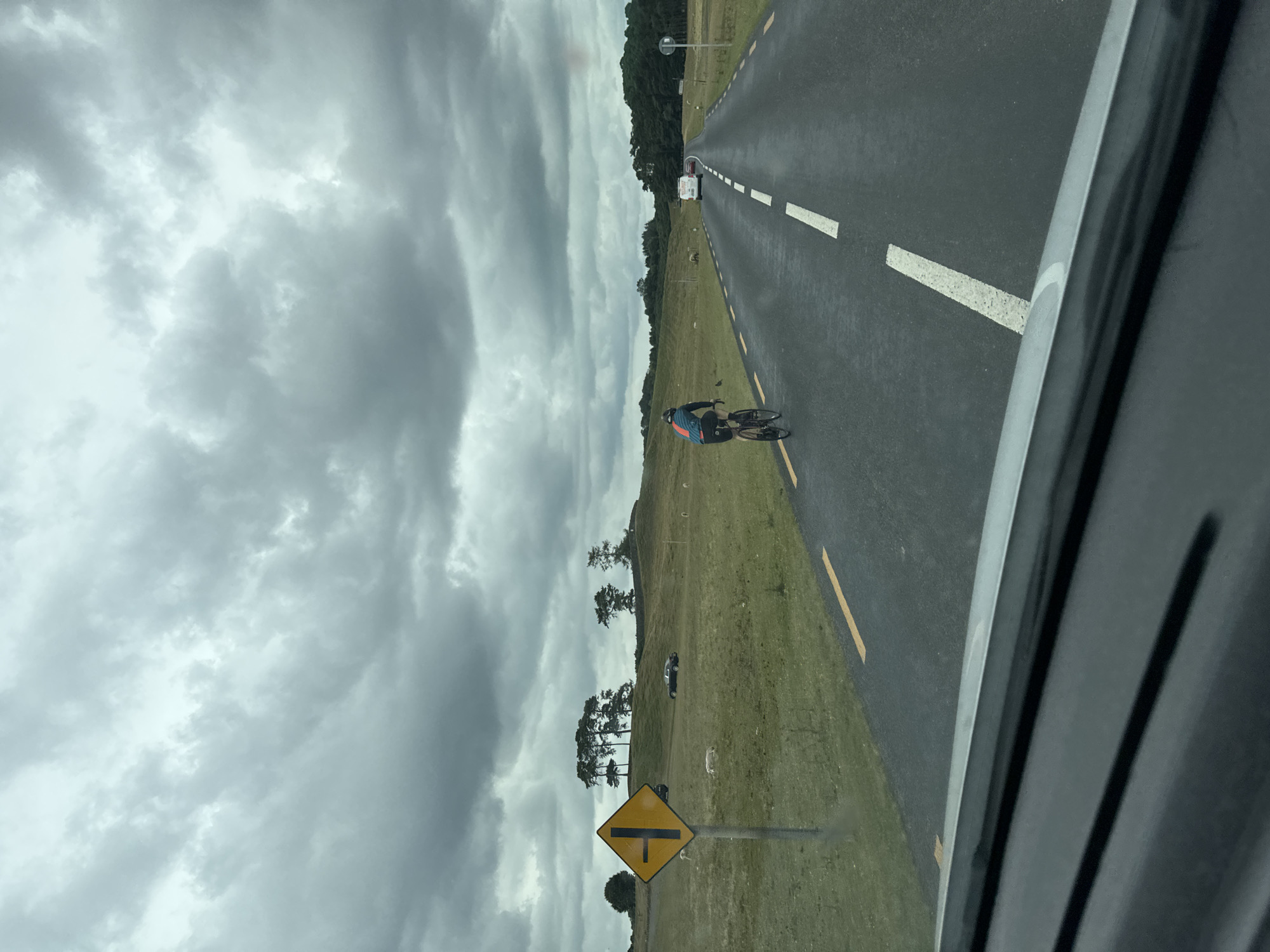 The image depicts a road scene with a cyclist in the foreground, riding on the right side of the road. The road is asphalt, with white dashed lines marking the lanes and yellow lines along the edges. The sky is overcast with dark, heavy clouds. To the left of the road, a yellow diamond-shaped sign with black markings indicates a junction ahead. A grassy field with some scattered sheep is visible beyond the sign. In the distance, there are trees and rolling hills. A few cars are also present on the road, further down. The image appears to be taken from a vehicle, with the dashboard visible at the bottom. There is no specific information in the image that can pinpoint the location to Newbridge, Ireland.
