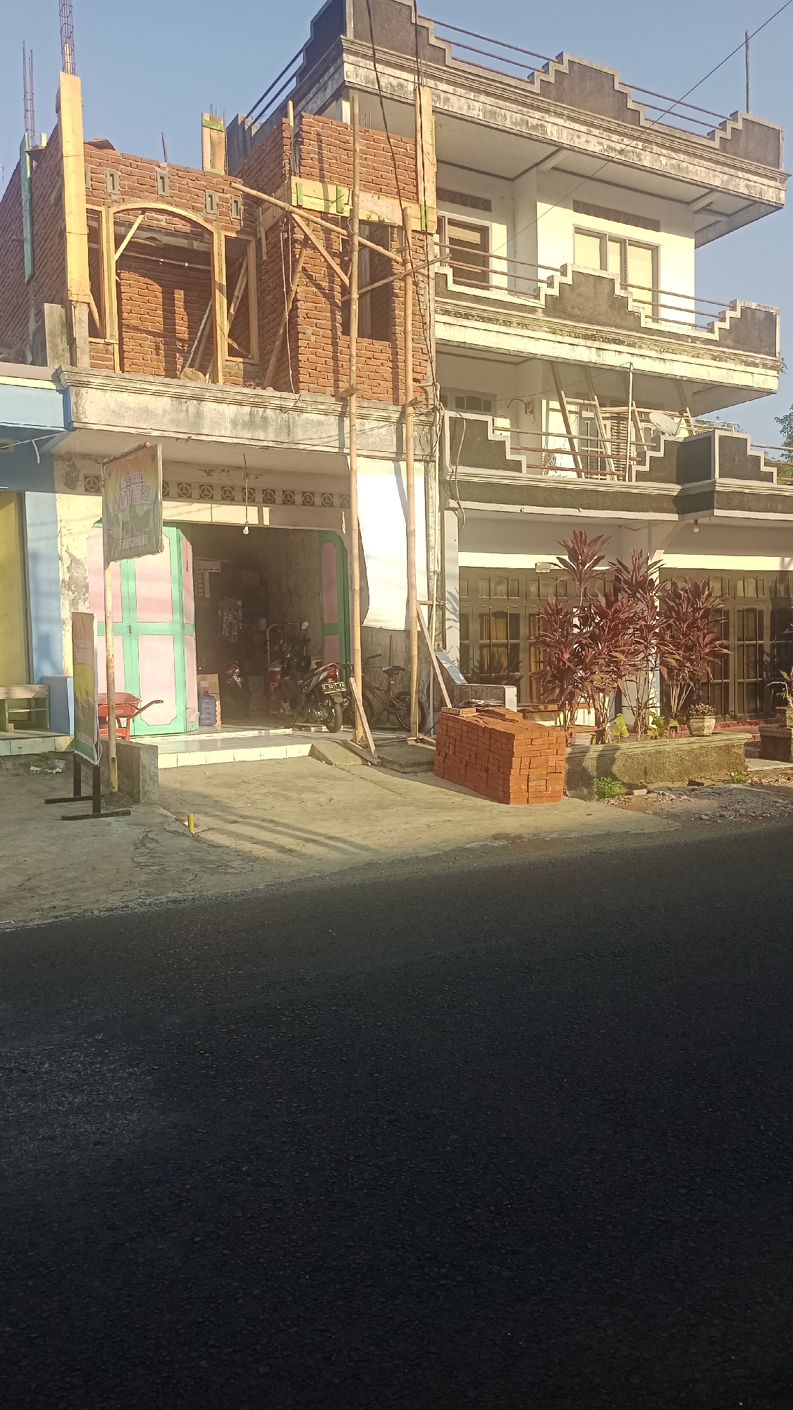 In Karyamandala, Indonesia, a building under construction is visible from a street level perspective. The construction site is in the foreground, with stacks of bricks placed on the ground. A motorcycle and bicycle are parked in front of a shop with open green and pink doors, indicating commercial activity. Scaffolding surrounds the unfinished brick wall of the building. The main structure appears to be a multi-story building with balconies and windows, partially completed. Red-leafed plants are situated in a planter box in front of the building. The sky is clear and blue, suggesting a sunny day.