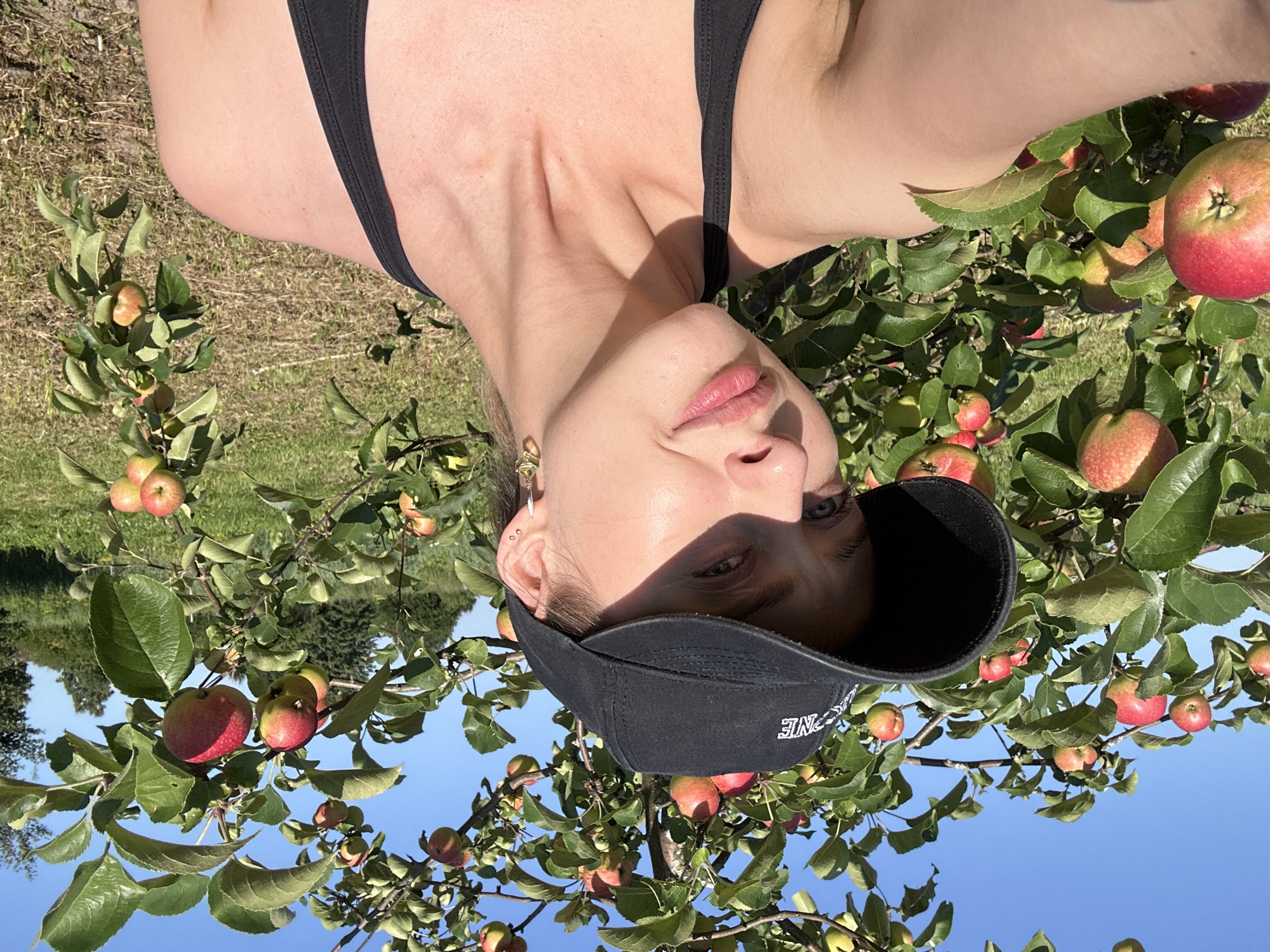 A young woman wearing a black baseball cap and a black tank top stands in front of an apple tree laden with ripe red apples. The woman is looking at the camera with a slight smile. The sun is shining brightly, casting shadows on her face. The background is a blur of green leaves and blue sky, with hints of trees and a field in the distance. The scene appears to be outdoors, possibly in an orchard or park. The image has a casual, summery feel.