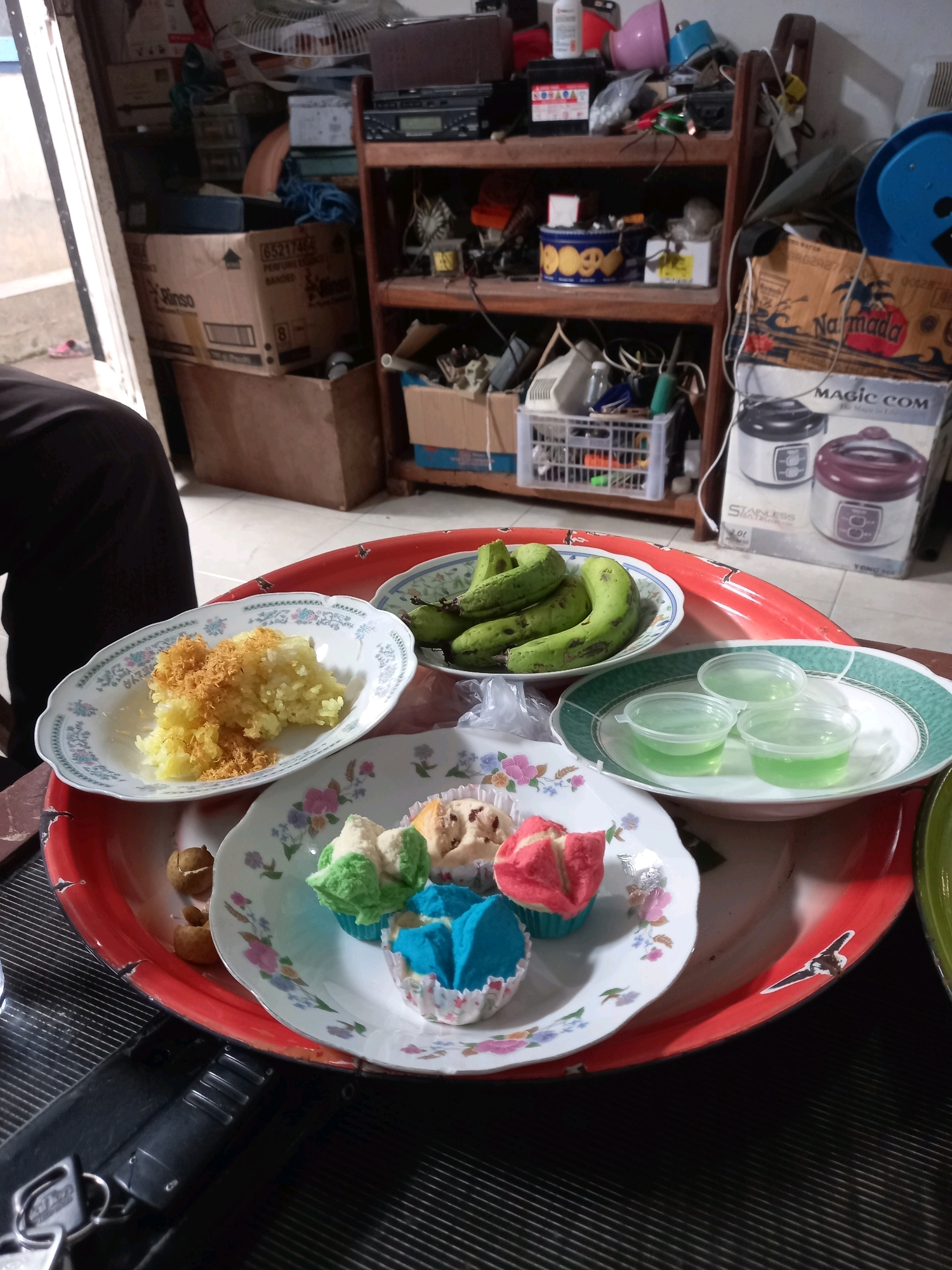 A high-angle, medium shot captures a red serving tray filled with Indonesian snacks and fruits. The tray is on a dark, textured surface, possibly a table. In the foreground, a person wearing black clothing is partially visible on the left. The tray holds three small white bowls. One contains a mound of yellow rice topped with shredded, light brown material. Another bowl holds several green bananas. A third bowl contains three small, clear plastic cups filled with a translucent green liquid, likely jelly or pudding. In the center of the tray, a floral-patterned plate displays an assortment of colorful cupcakes and some small, brown, nut-like objects. The cupcakes are shaped like flowers and colored pink, blue, and green, with one patterned with dark specks. Behind the tray, a shelf unit is visible, filled with various items including boxes, electronics, and household supplies. The background suggests an indoor setting, possibly a storage area or a kitchen in a home. The overall scene depicts a collection of food items ready for consumption.