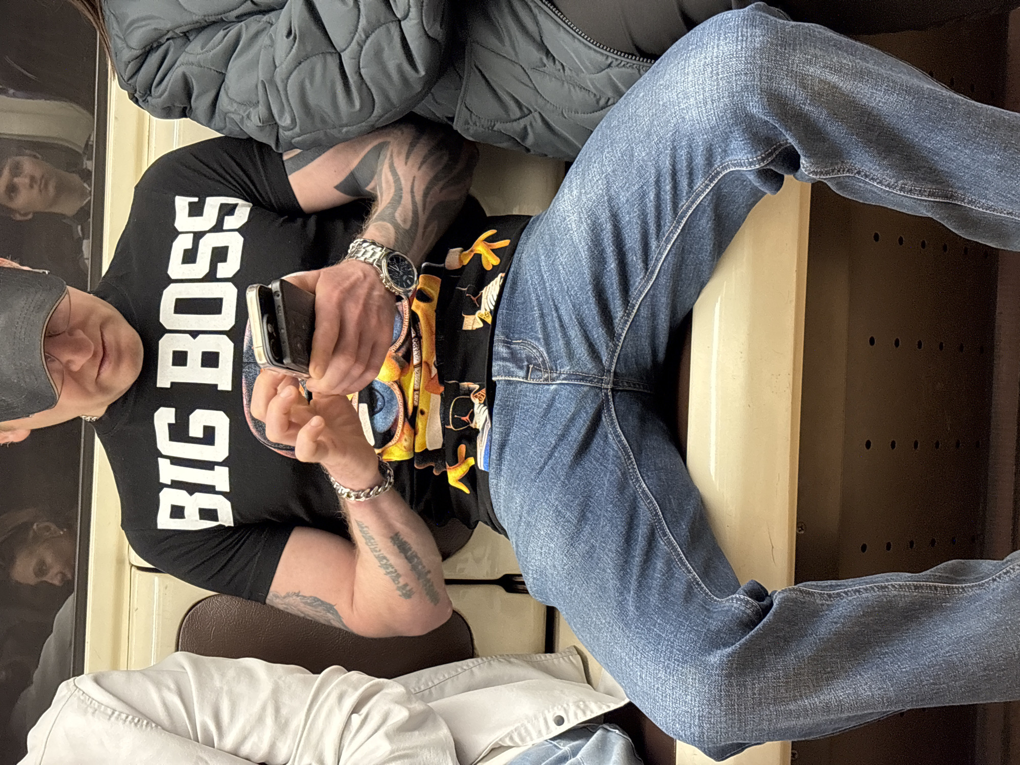 This image captures a man seated in what appears to be a public transport vehicle, consistent with a setting in Saint Petersburg, Russia. The man is wearing a black t-shirt with the white text "BIG BOSS" prominently displayed across the chest. He also wears blue jeans, a black baseball cap, a silver watch on his left wrist, and a silver chain bracelet on his right wrist. Both of his forearms are heavily tattooed, with tribal-like patterns visible on his left arm and script on his right.
He is holding a smartphone with both hands, looking down at the screen, indicating he is engaged with the device. The interior of the vehicle features beige and brown surfaces, including a backrest and what looks like a window or partition with a reflective quality. The indistinct reflection of a person's face can be seen in the dark, reflective surface to the man's left.
Above the man, the torso and arm of another person are visible, dressed in a dark grey quilted jacket. Below him, the lower body and legs of another passenger, wearing light-colored trousers, are partially visible. The scene depicts a common daily activity of individuals commuting and using their personal electronic devices in transit. The lighting suggests an indoor environment, but the time of day or weather outside is not discernible.