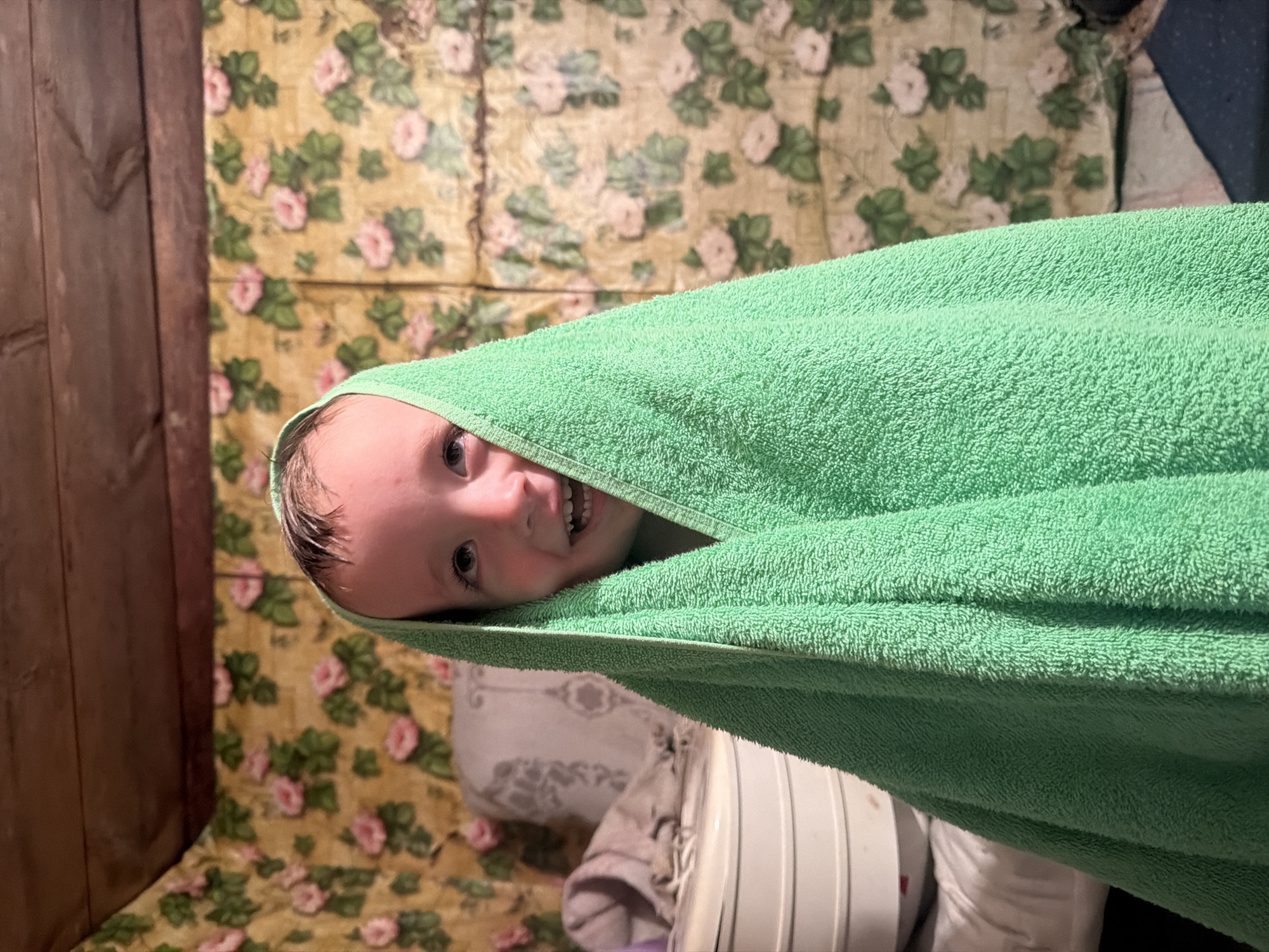 A young child, appearing to be a boy, is wrapped in a bright green towel. The towel covers most of his body, with only his face visible. He has short, dark hair that appears wet and is looking upwards with a wide, toothy smile. The background of the image consists of floral wallpaper with pink and green roses on a yellow background, and wooden planks at the top of the frame, suggesting an indoor setting. To the left of the child, there is a stack of white containers and a pillow with a grey pattern. The scene has a casual, home-like atmosphere. The location of Красный Маяк, Russia is not explicitly evident from the image itself and would require external information to confirm.